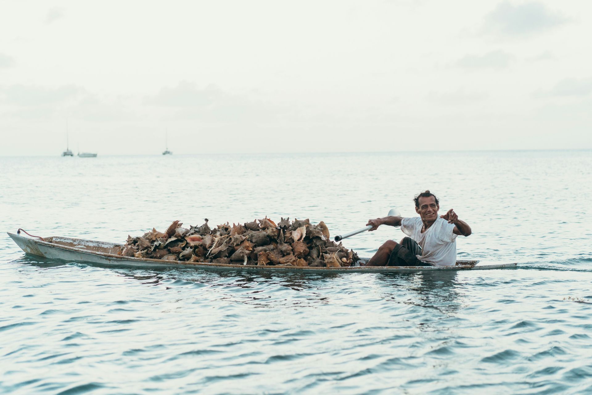 A man pulls a small boat laden with piled objects through calm water. The sky is overcast in Caye Caulker, Belize.