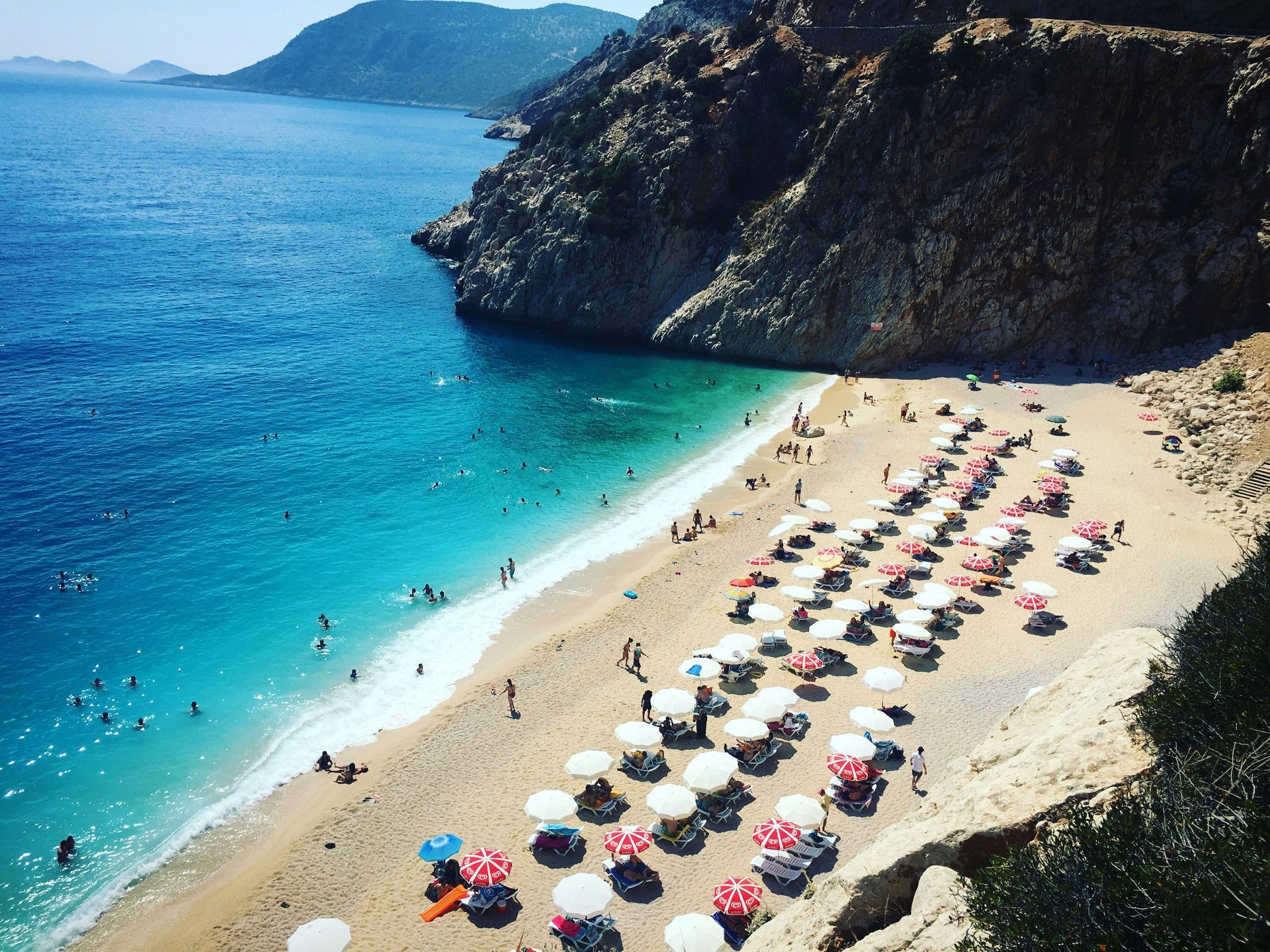 Beach with turquoise water and white umbrellas and people swimming, nestled against a cliff in Kaş, Turkey.