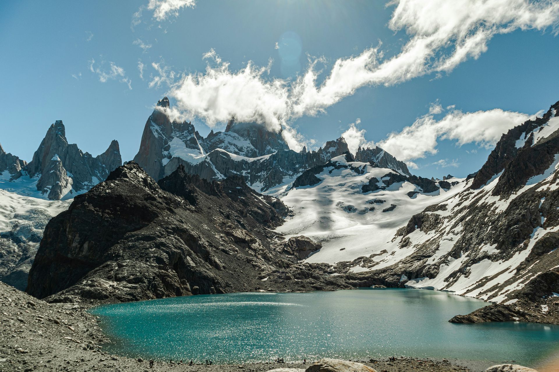 Lake beneath snow-capped mountains, turquoise water reflecting a blue sky with clouds in El Chaltén, Argentina.