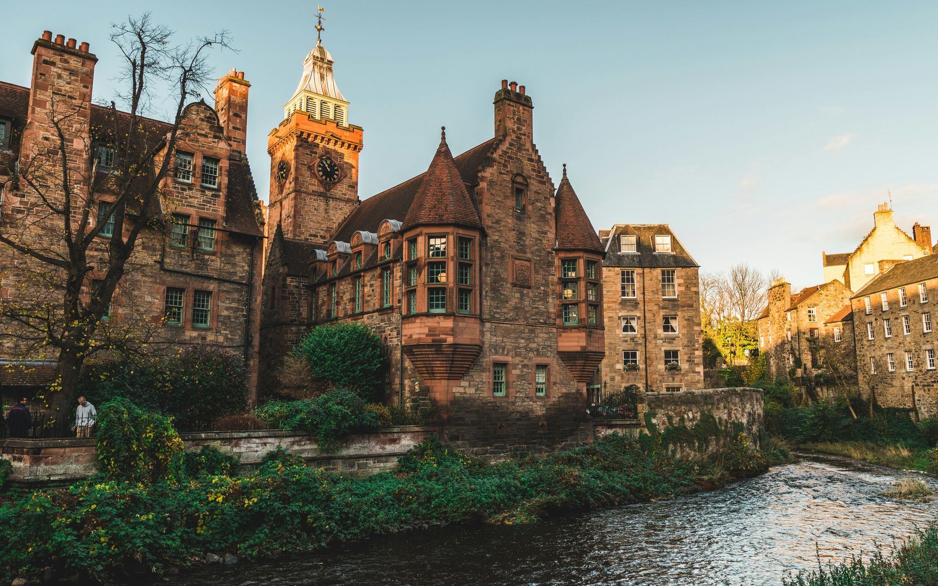 Stone buildings with a clock tower, next to a stream lined with greenery in Edinburgh, Scotland.
