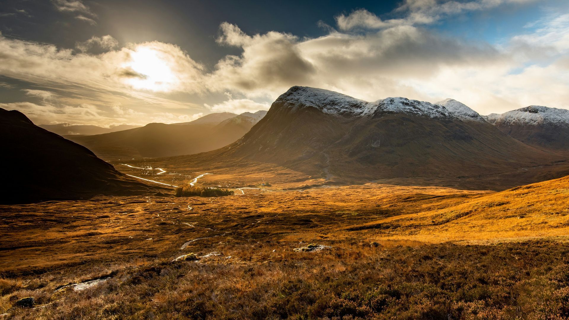 Sunlit mountain range with snow, overlooking a valley of brown brush and a town, with cloudy sky in Glencoe, Scotland.