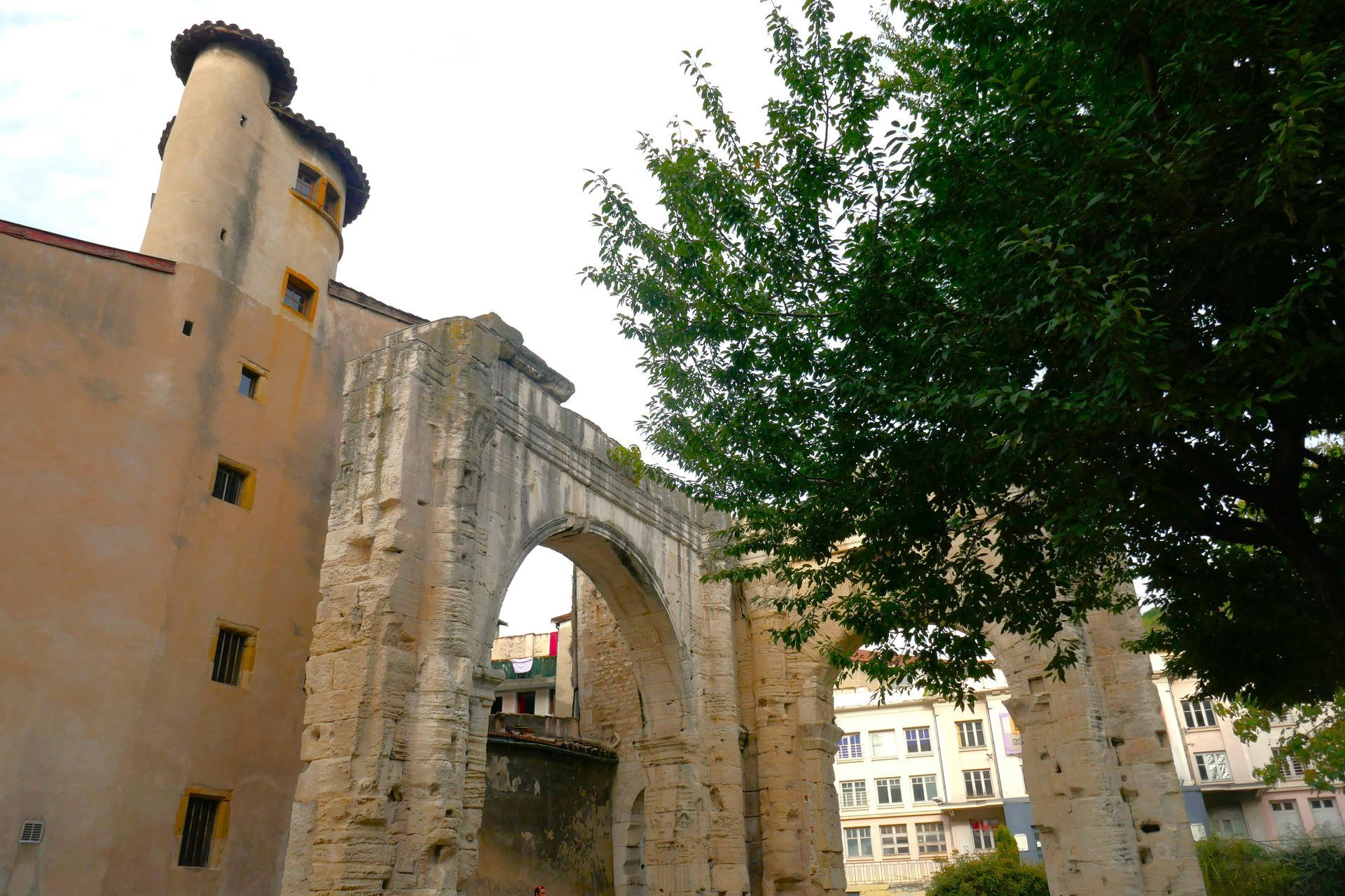Stone ruins with an archway and a round tower, set against a backdrop of buildings and a tree.