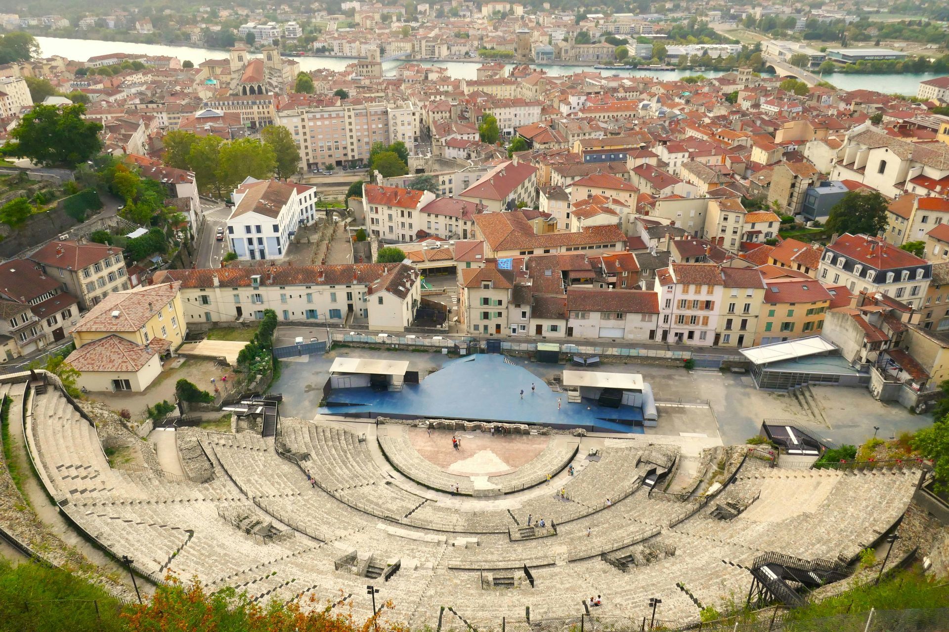 Aerial view: Roman theater with surrounding city buildings. Blue stage setup in the center. Brown roofs, green trees.