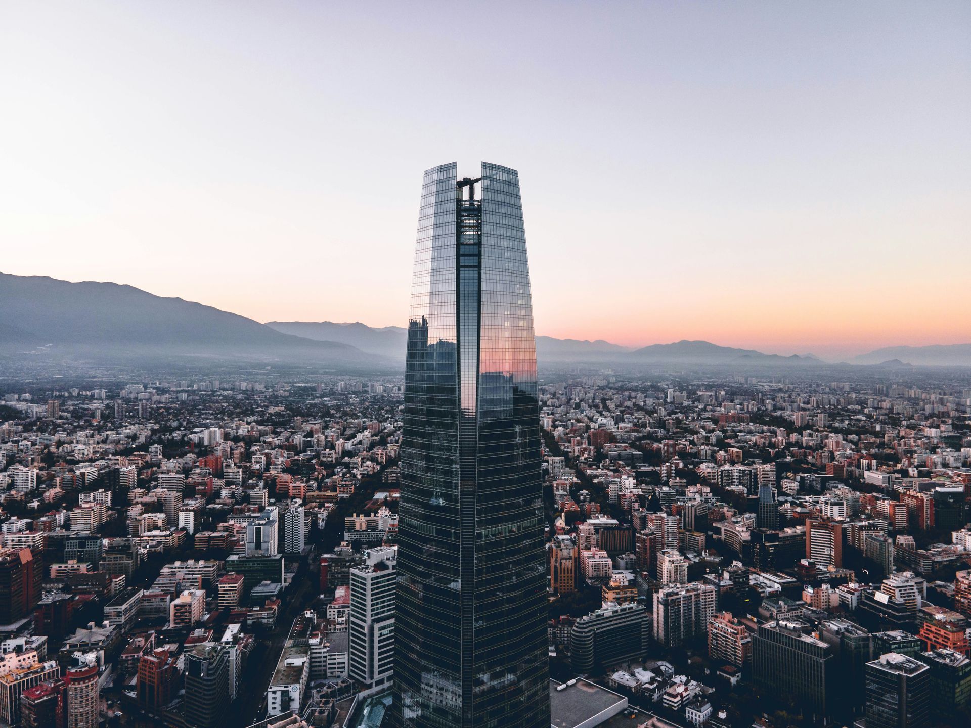 Skyline view with a tall, mirrored skyscraper in the center, surrounded by city buildings, at sunset in Santiago, Chile.
