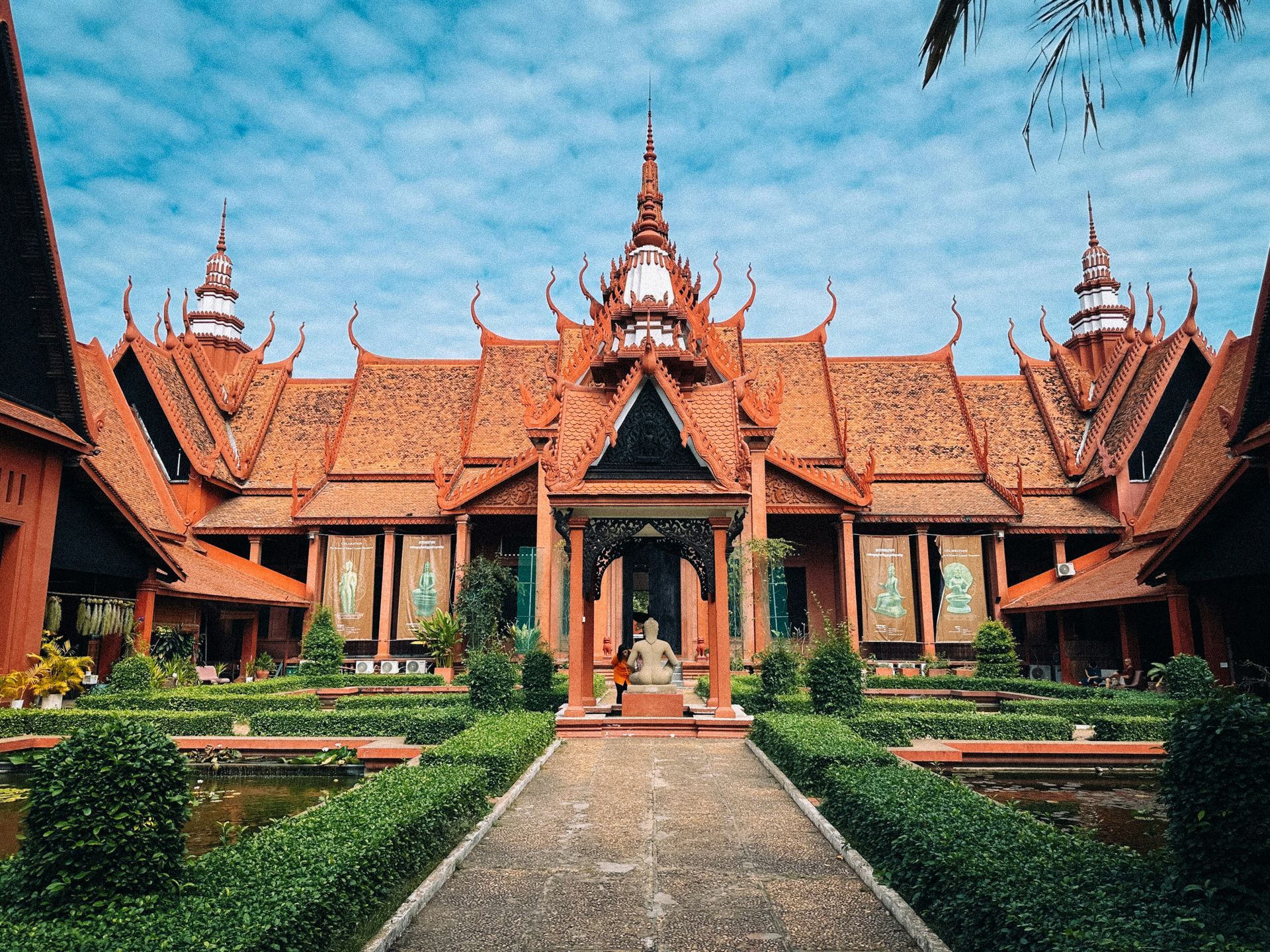 National Museum of Cambodia in Phnom Penh, red-roofed building with ornate architecture, lush gardens, and a statue.