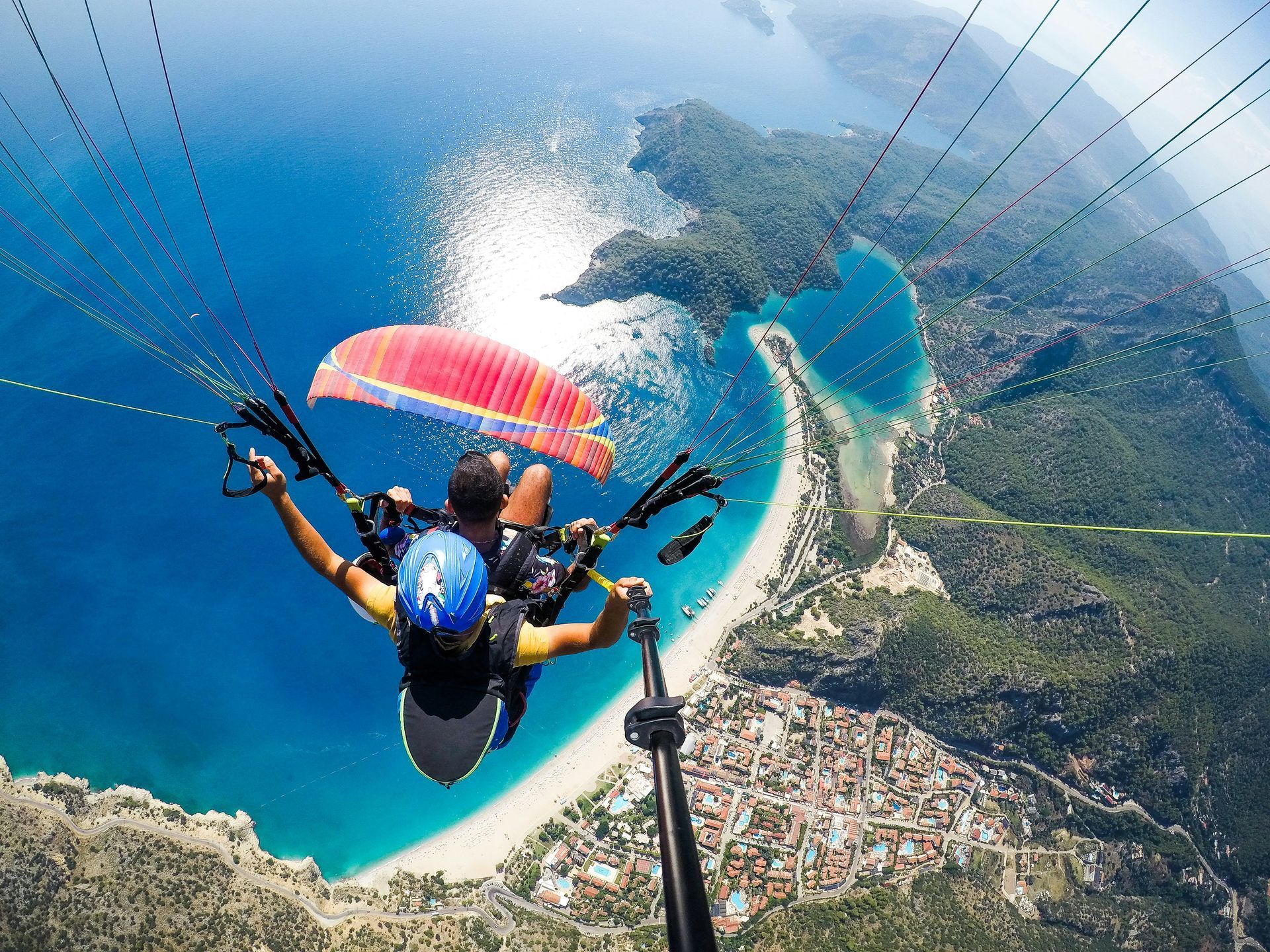 Paragliders soar over a turquoise bay and sandy beach, taking a selfie in Fethiye, Turkey.