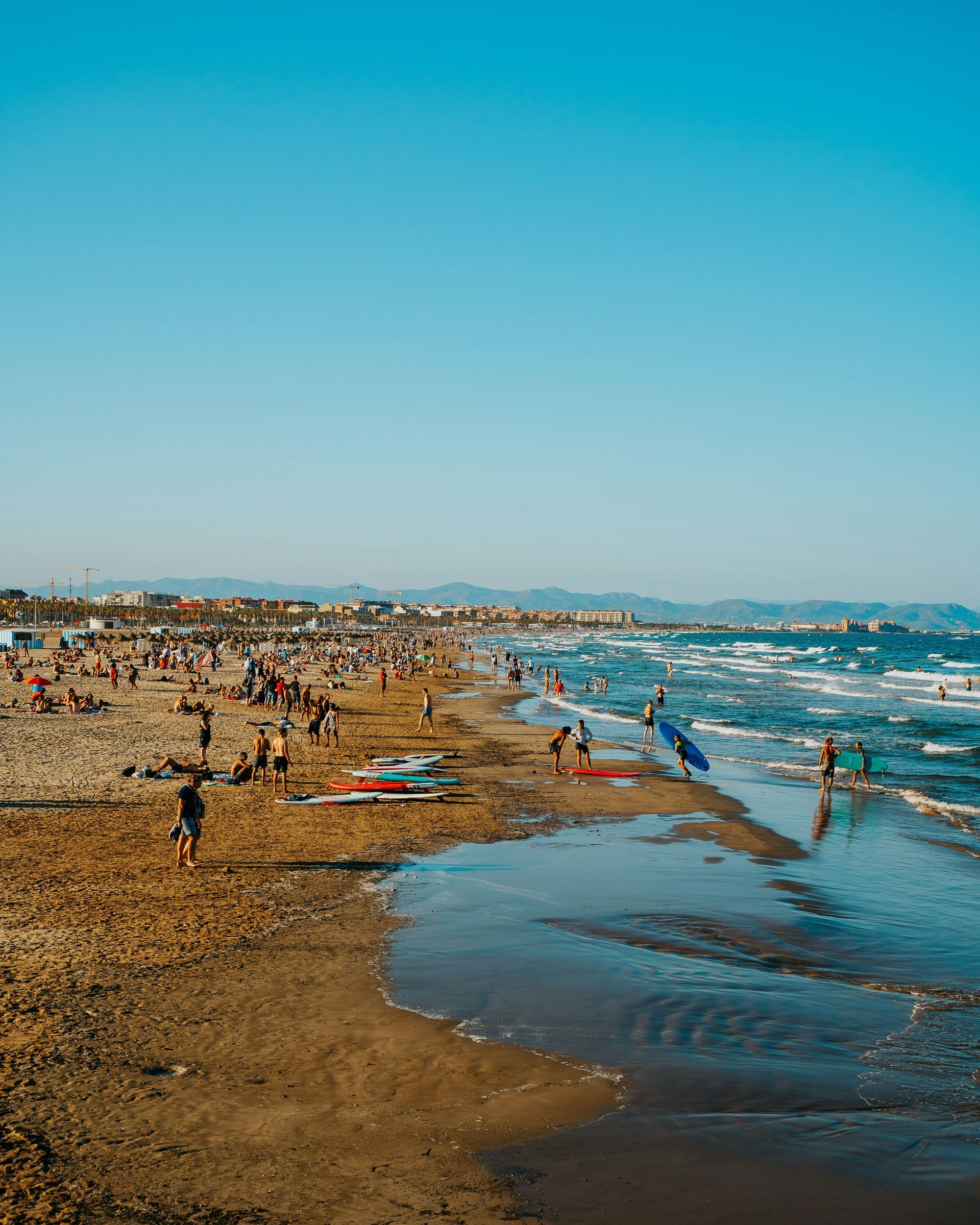 Beach scene with many people, blue sky, waves, and some surfers on a sunny day in Valencia, Spain.