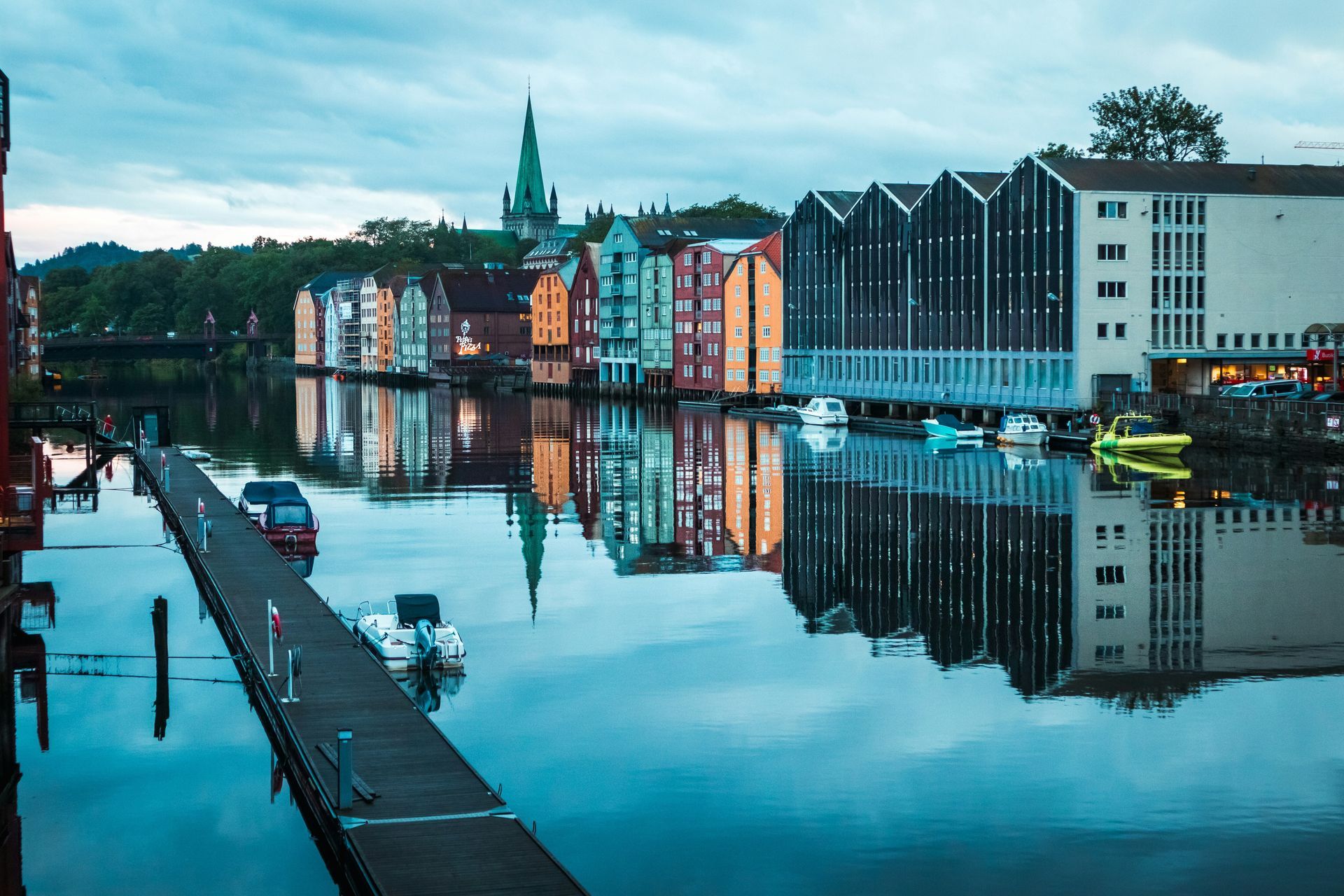 Colorful buildings line a calm river, reflecting in the water; a pier extends toward the buildings in Trondheim, Norway.