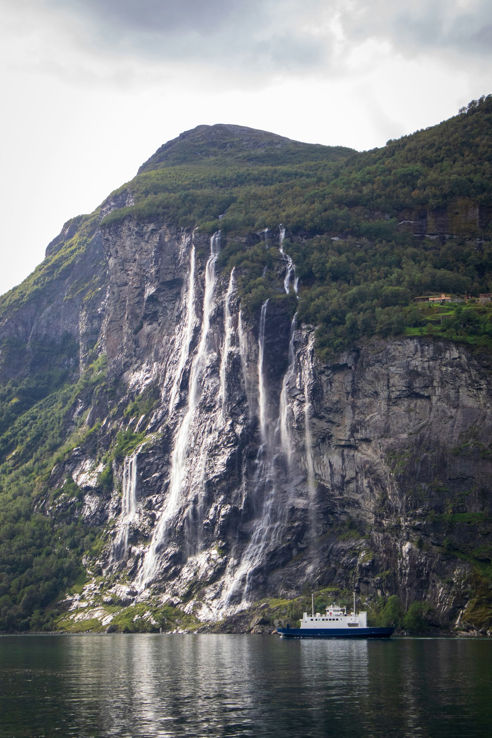 Waterfall cascading down a rocky mountain into the Geirangerfjord; ferry boat travels at base in Norway.