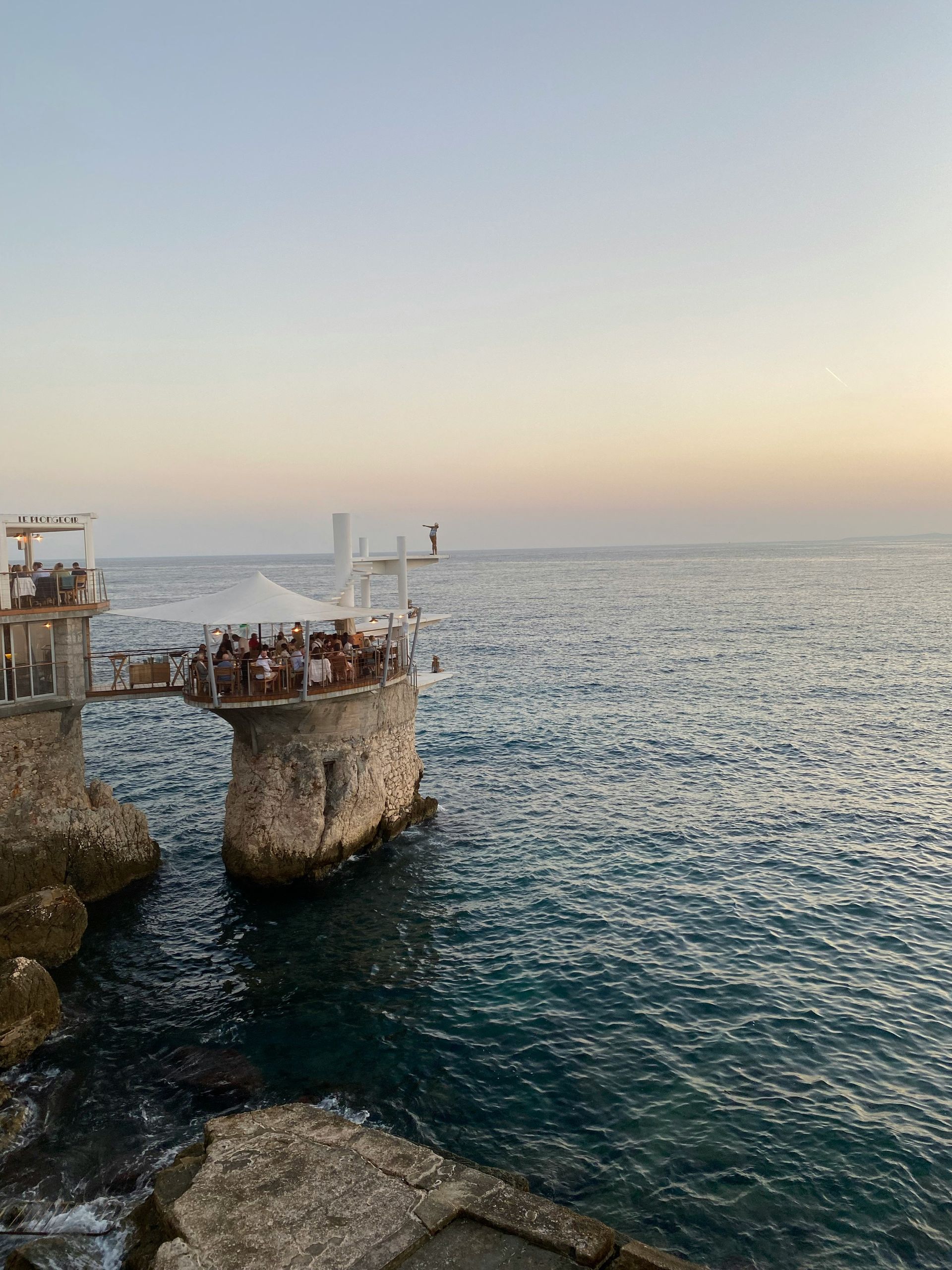 A group of people are eating on top of a cliff in the middle of the ocean at the restaurant Le Plongeoir in Nice, France.