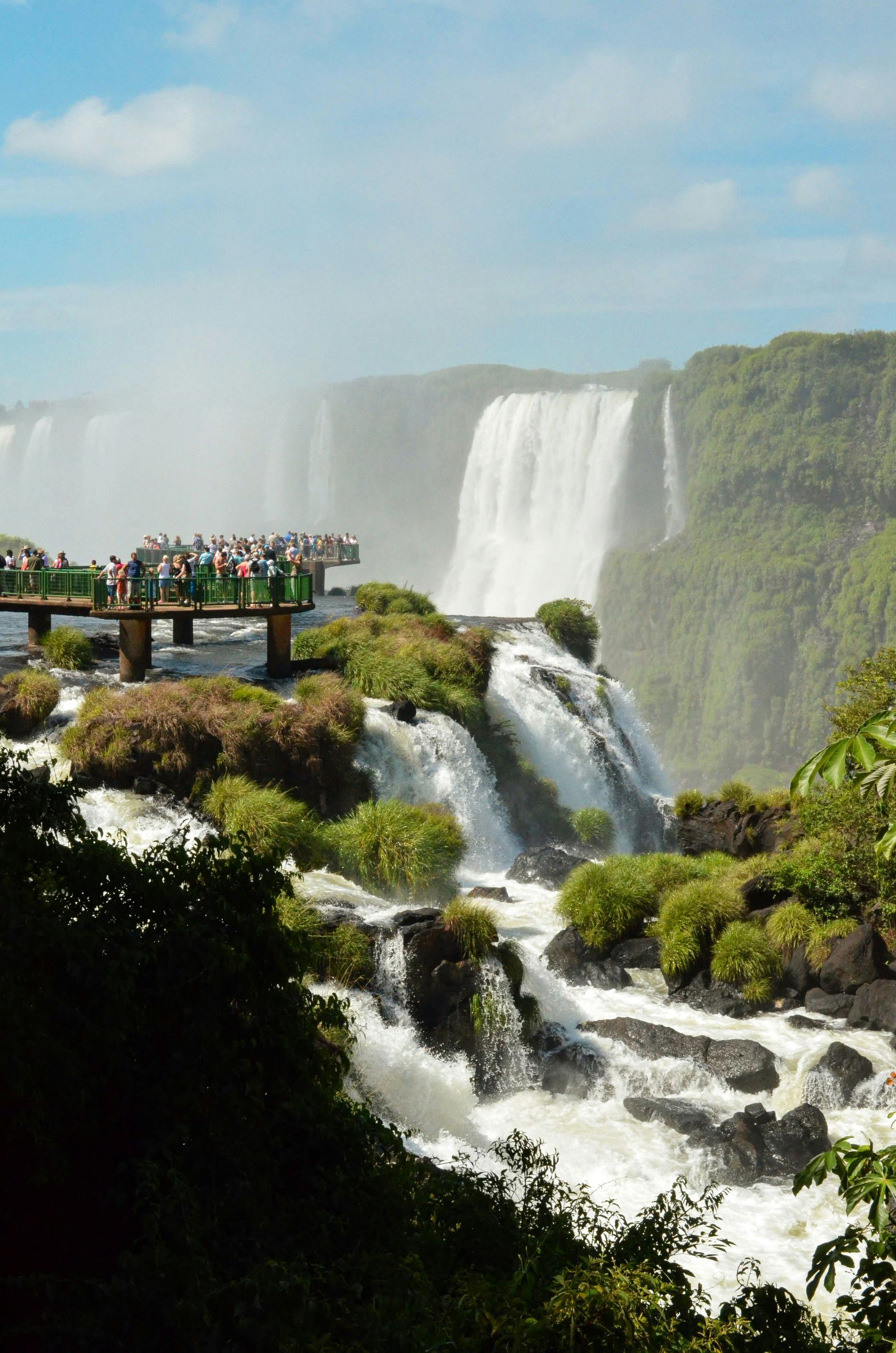 Iguazu Falls, Argentina: Tourists on a viewing platform, waterfalls cascading over rocks, lush greenery under blue sky.