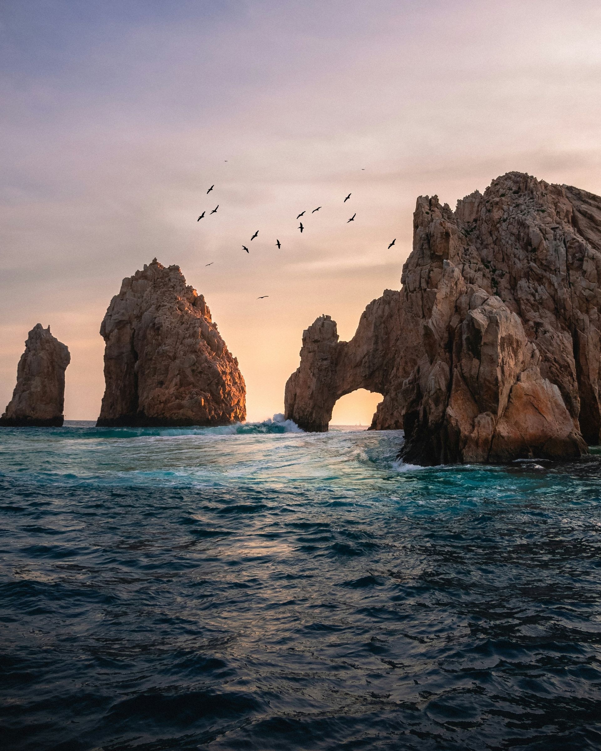 Rocky cliffs jutting from a turquoise ocean at sunset, with birds flying overhead in Cabo San Lucas, Mexico.