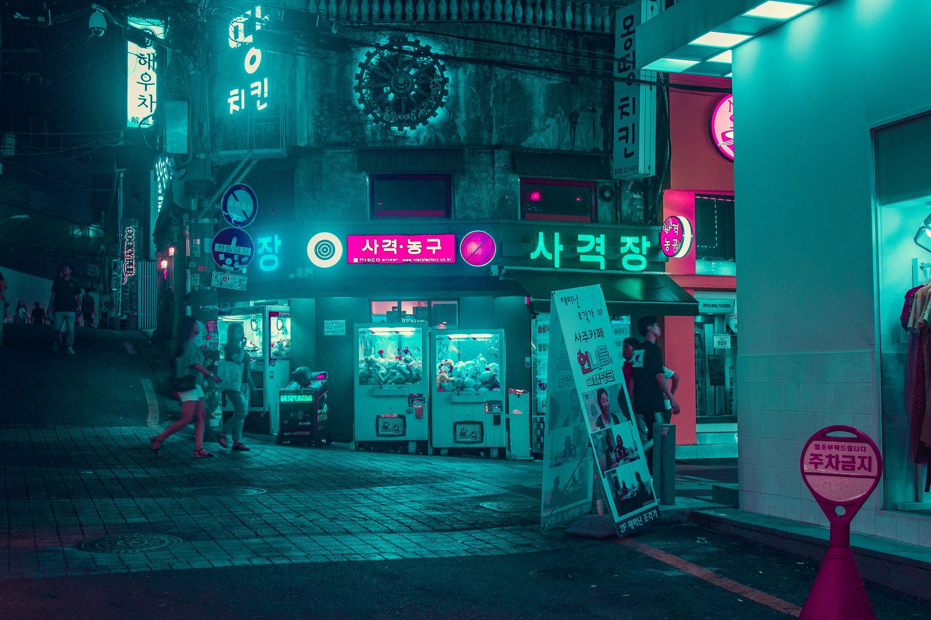 Night scene in South Korea. Neon lights illuminate buildings and street. Claw machines, signs, and pedestrians present.