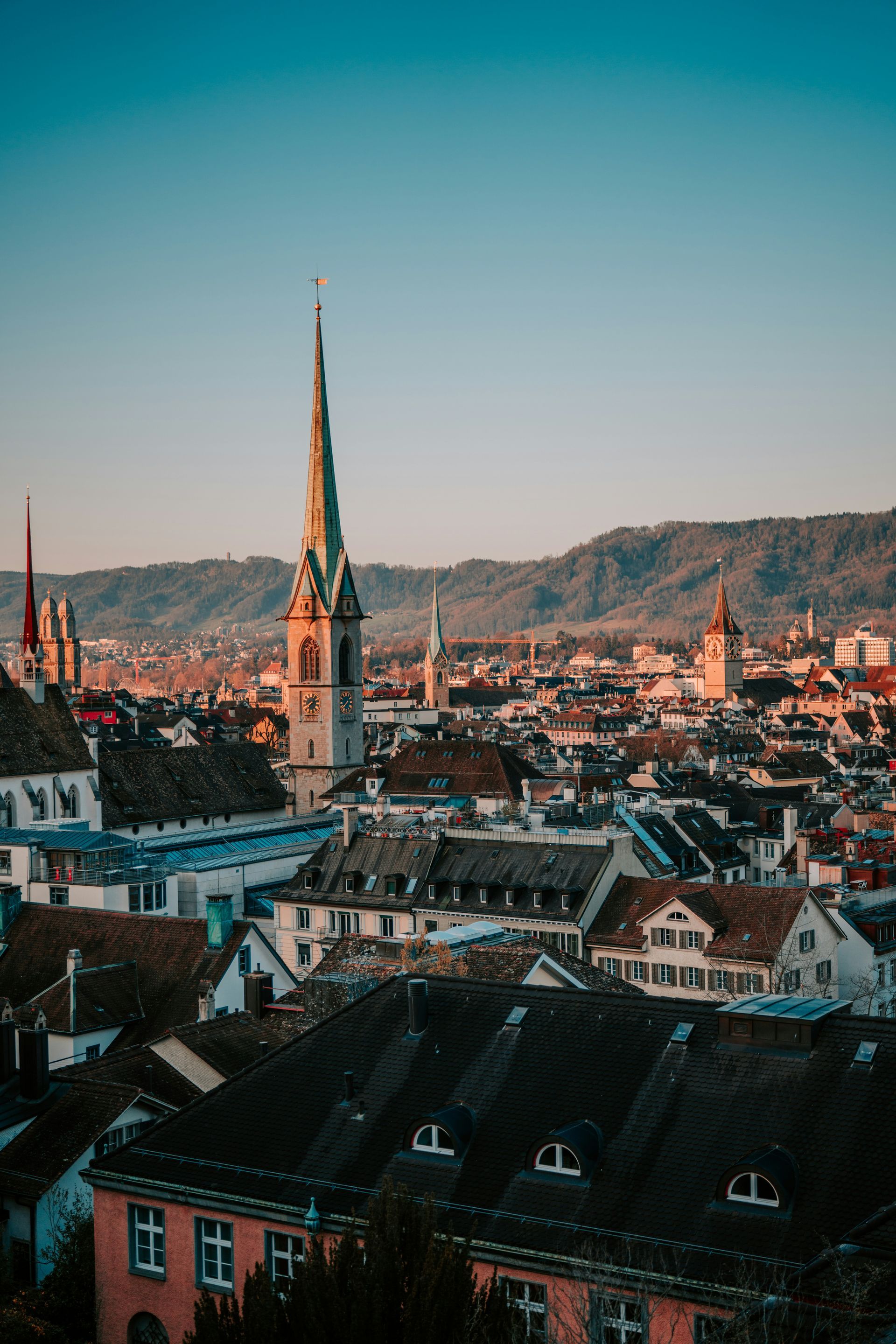 Cityscape of Zurich, Switzerland, with church steeple, rooftops, and mountains under a blue sky.