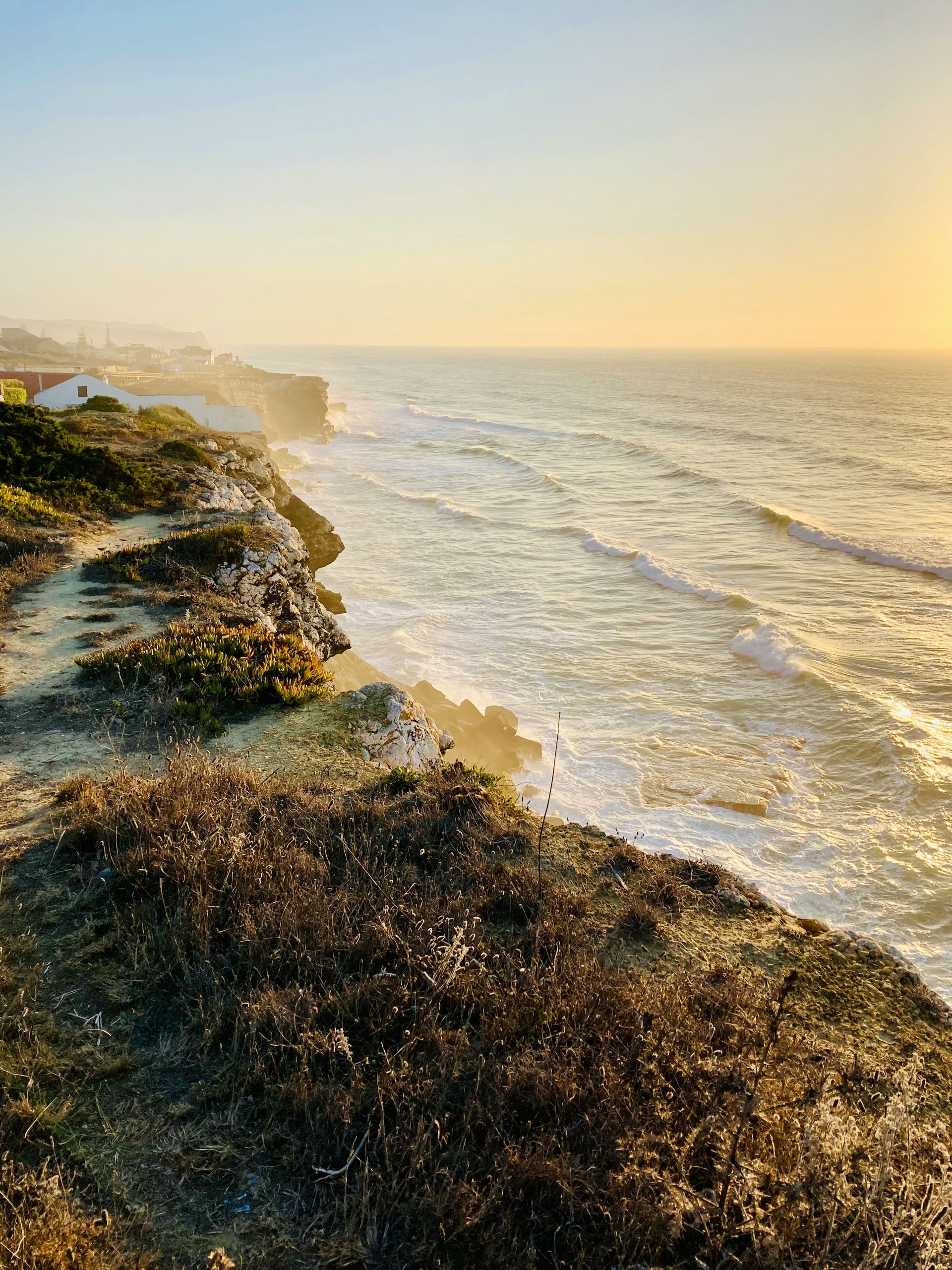 Coastal cliff overlooking the ocean during golden hour; sunlit waves crash below in Azenhas do Mar, Portugal.