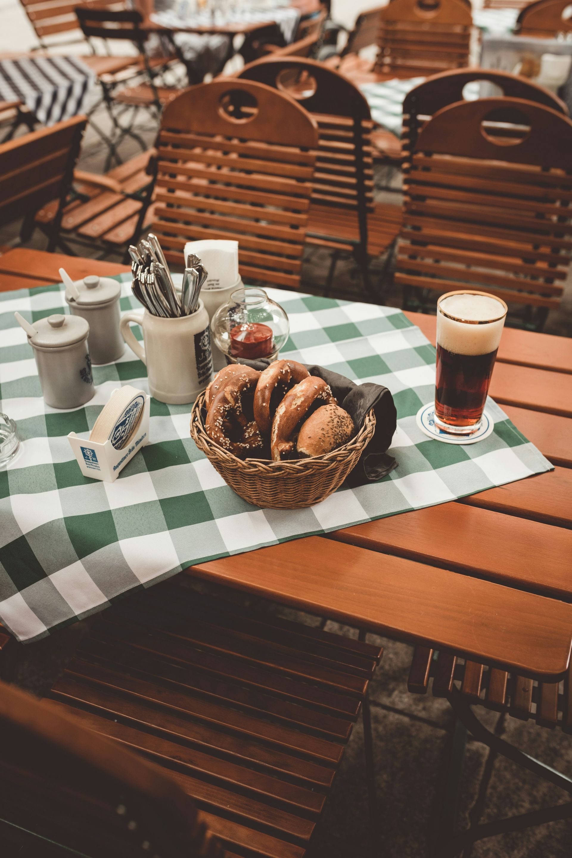 Outdoor cafe table with a basket of pretzels, beer, and condiments, and a green checkered tablecloth in Germany.