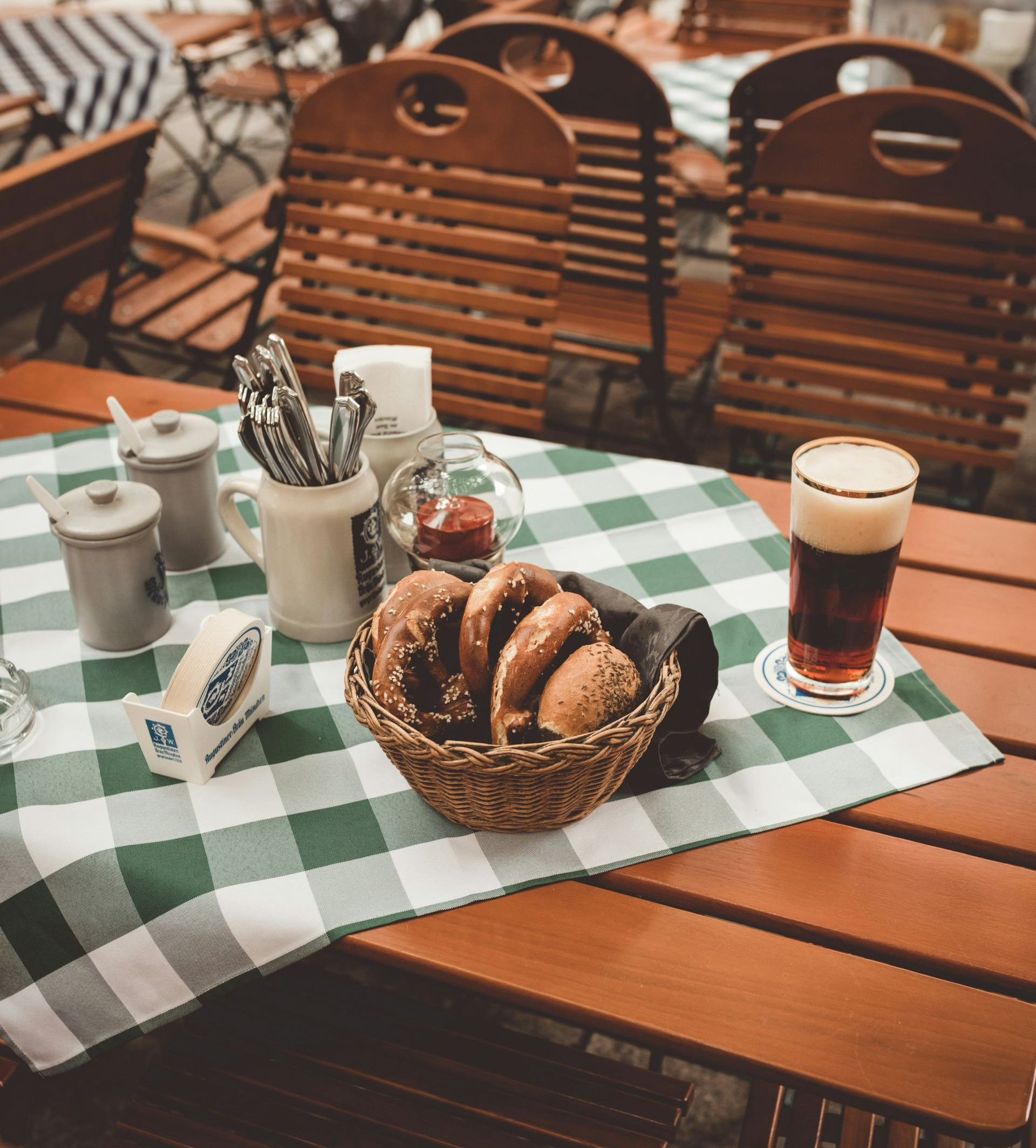Outdoor cafe table with a basket of pretzels, beer, and condiments, and a green checkered tablecloth in Germany.