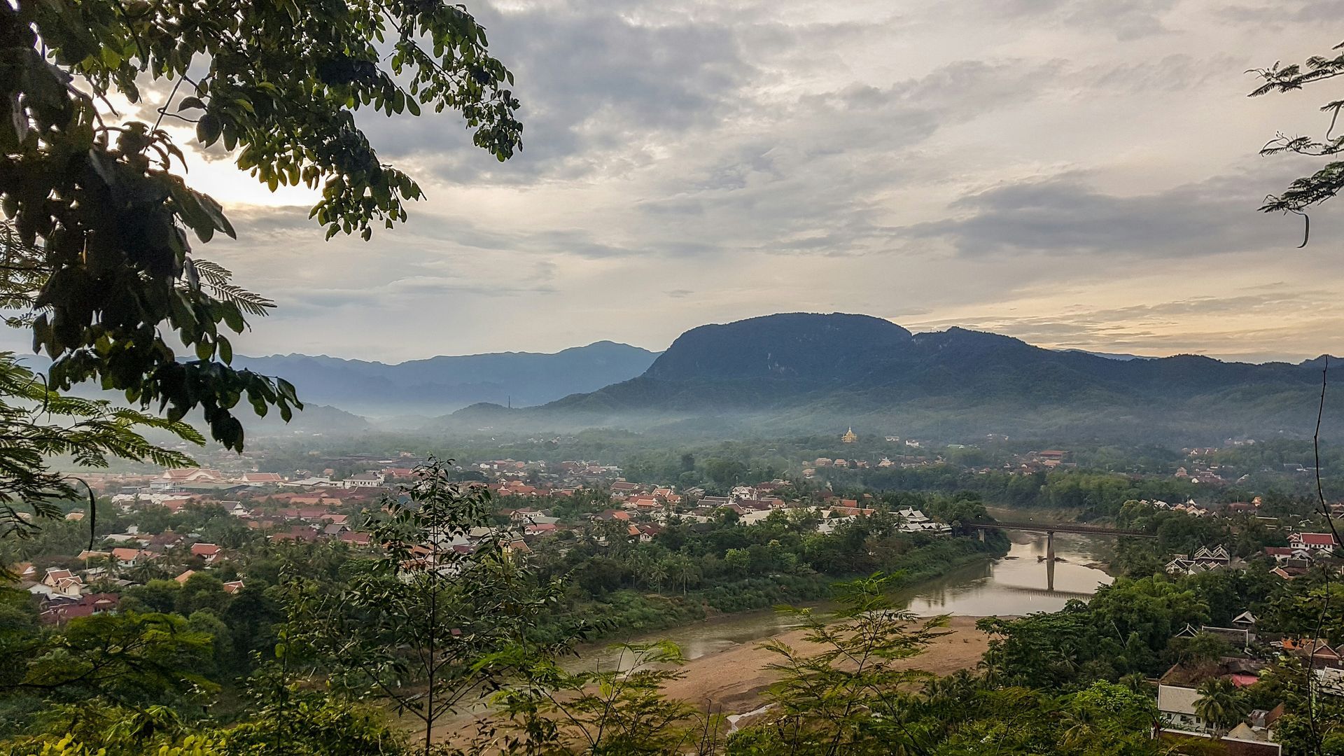 Scenic view of Luang Prabang, Laos, with misty mountains, a river, and buildings nestled in the valley.