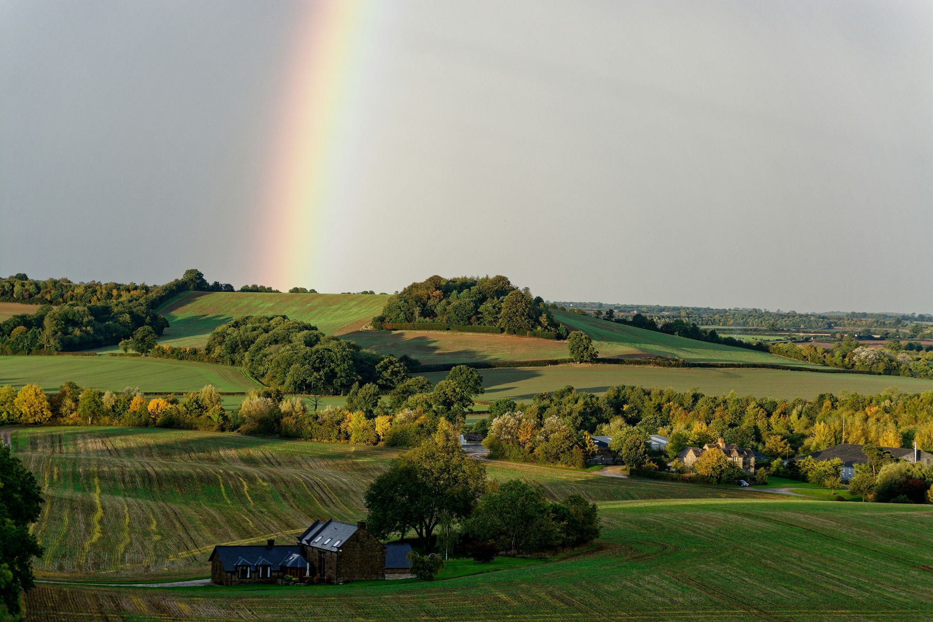 Rainbow arcs over rolling green hills with scattered trees and small buildings in the Cotswolds, England.