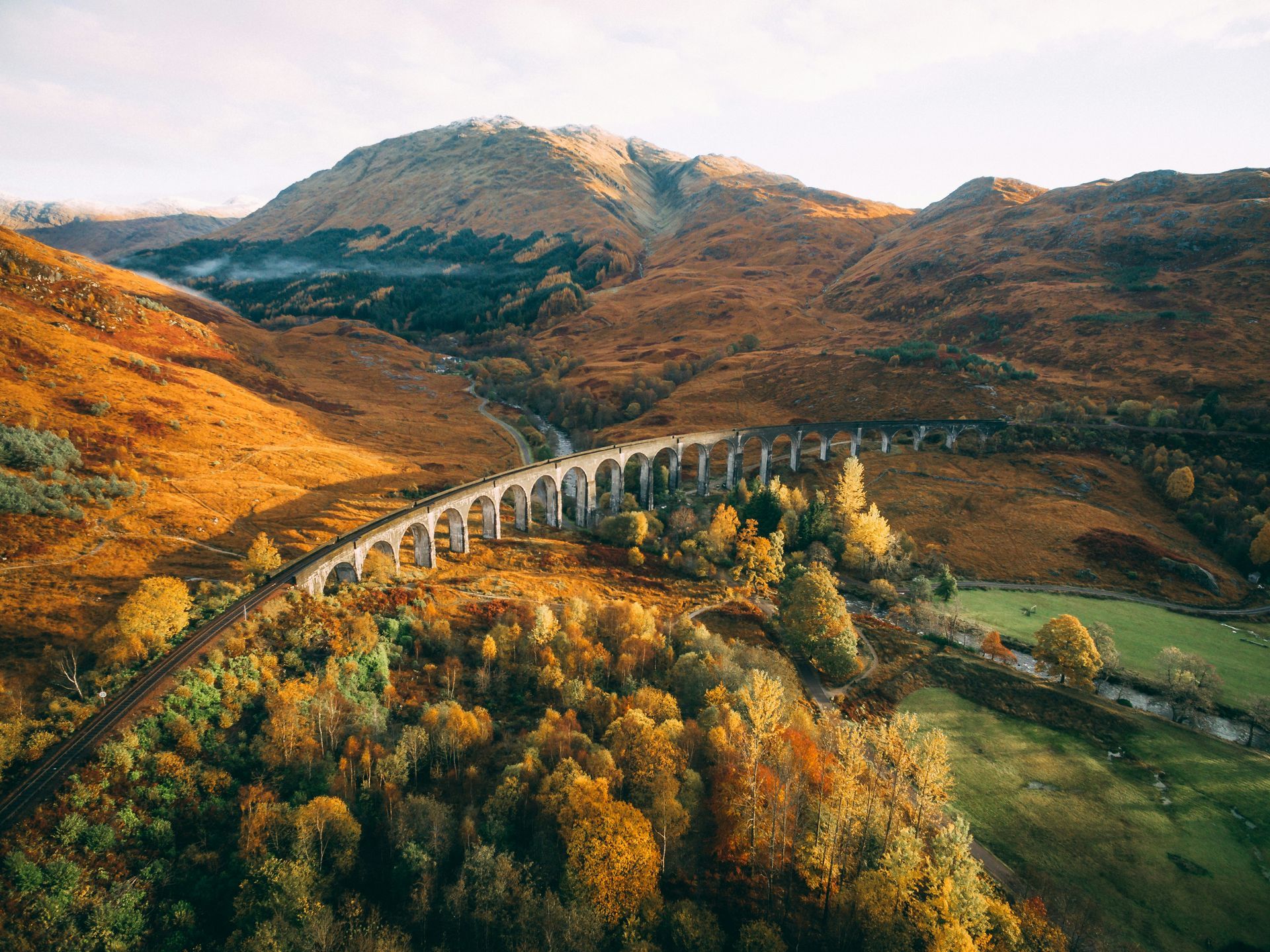 The Glenfinnan Viaduct railway viaduct arches over a valley in autumn, surrounded by golden trees and mountains in Scotland.