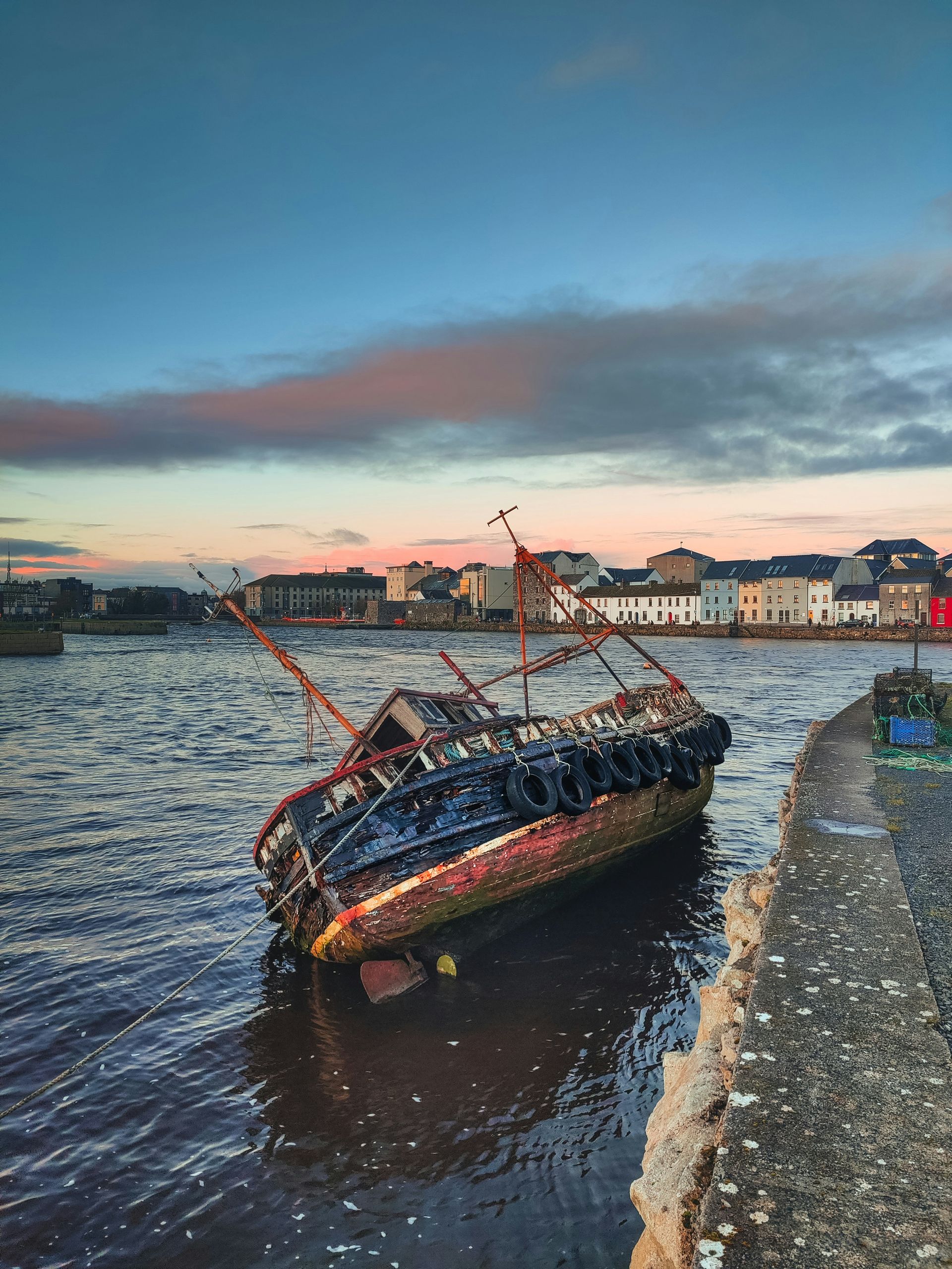 Decaying boat in water, near a stone pier, with a town in the background at sunset in Galway, Ireland.