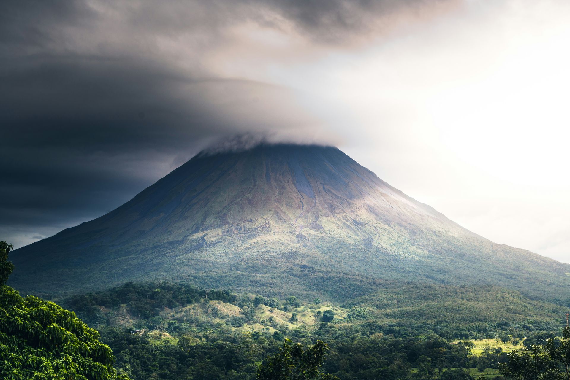 The Arenal Volcano summit shrouded in clouds; lush green forests at its base. Dramatic sky and sunlight in Costa Rica.
