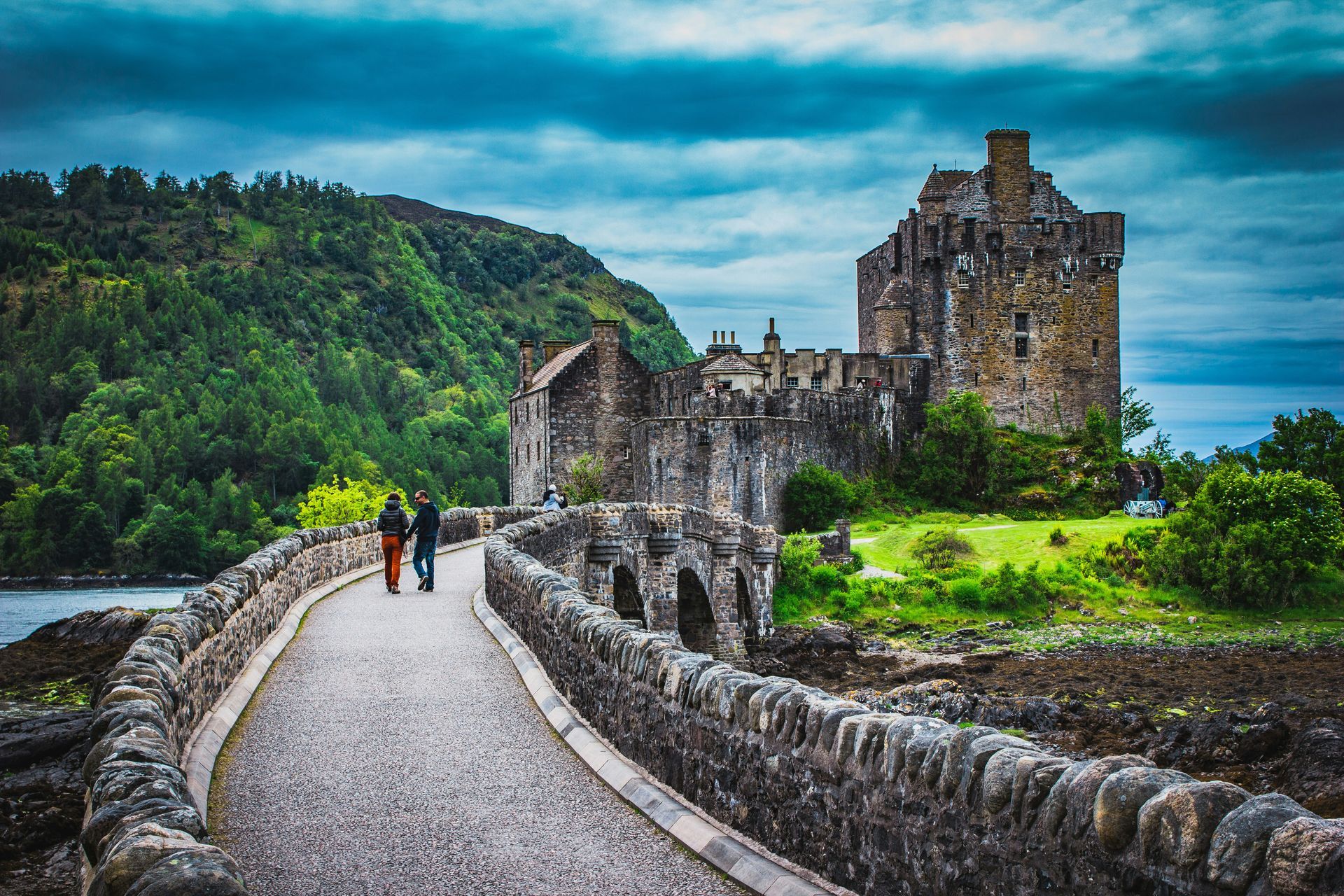 The Eilean Donan Castle with a bridge, two people walk on bridge. Lush green hills and blue sea in Scotland.