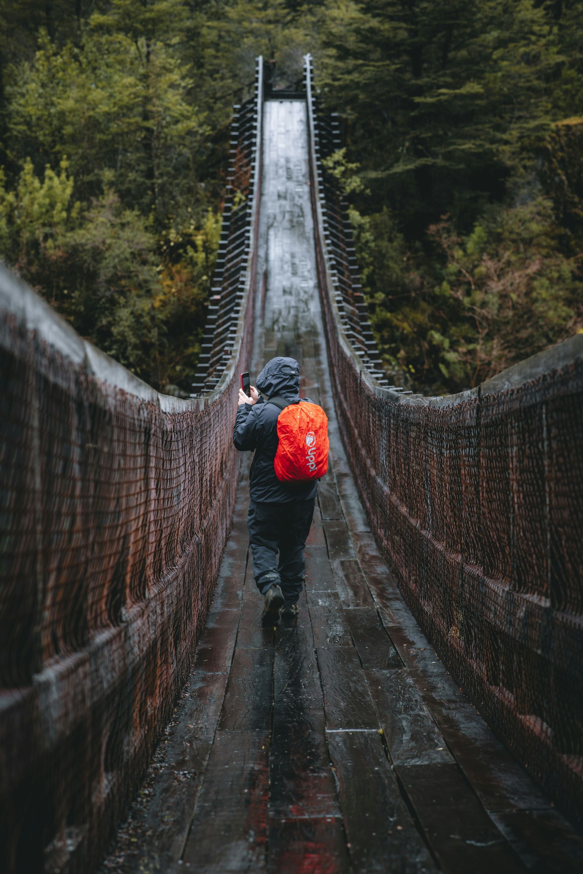 Person with backpack walks across a weathered suspension bridge in a rainy forest in Aysén, Chile.