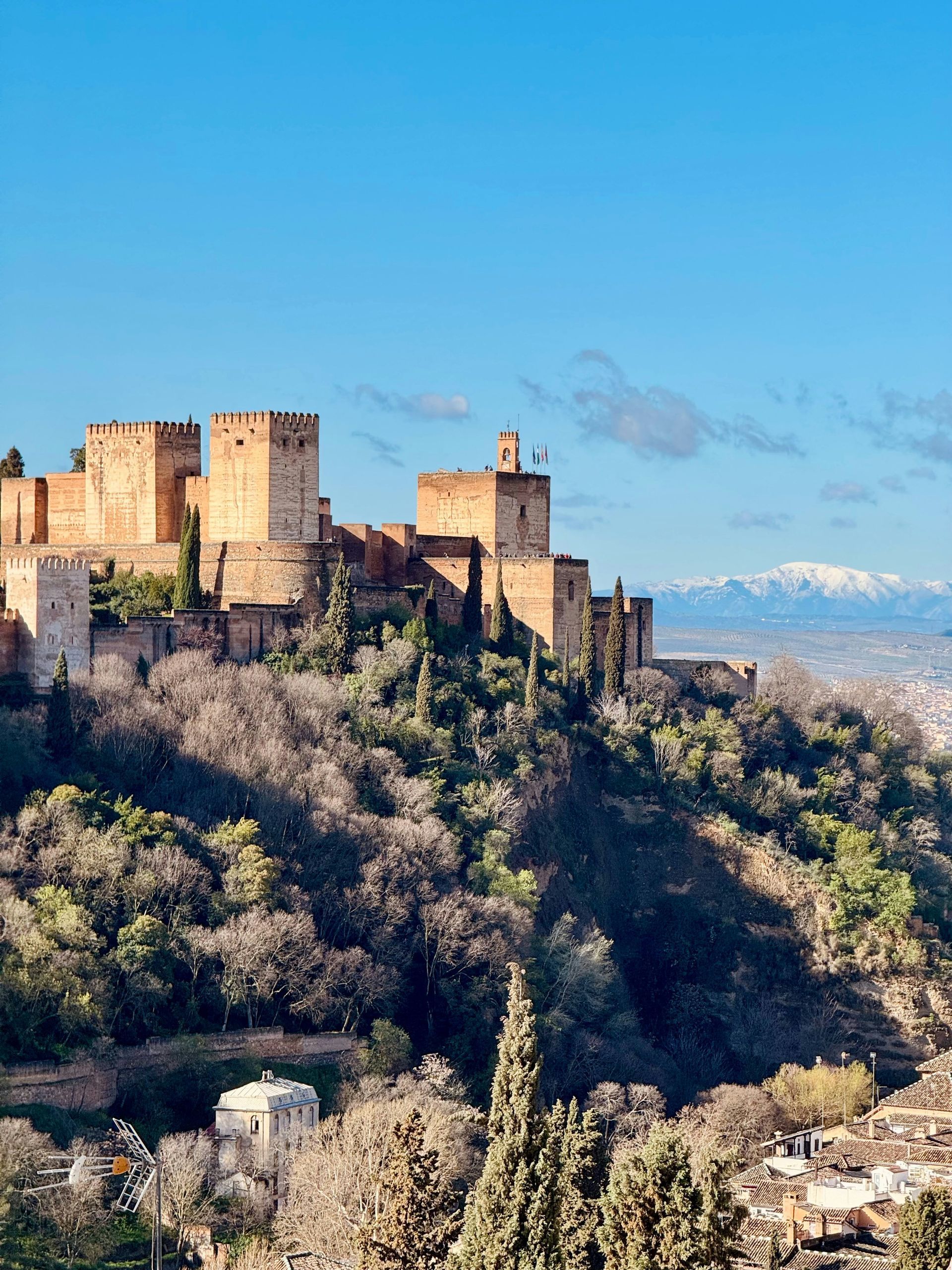 Granada's Alhambra palace rises on a hill, golden against a blue sky; snow-capped mountains in background.