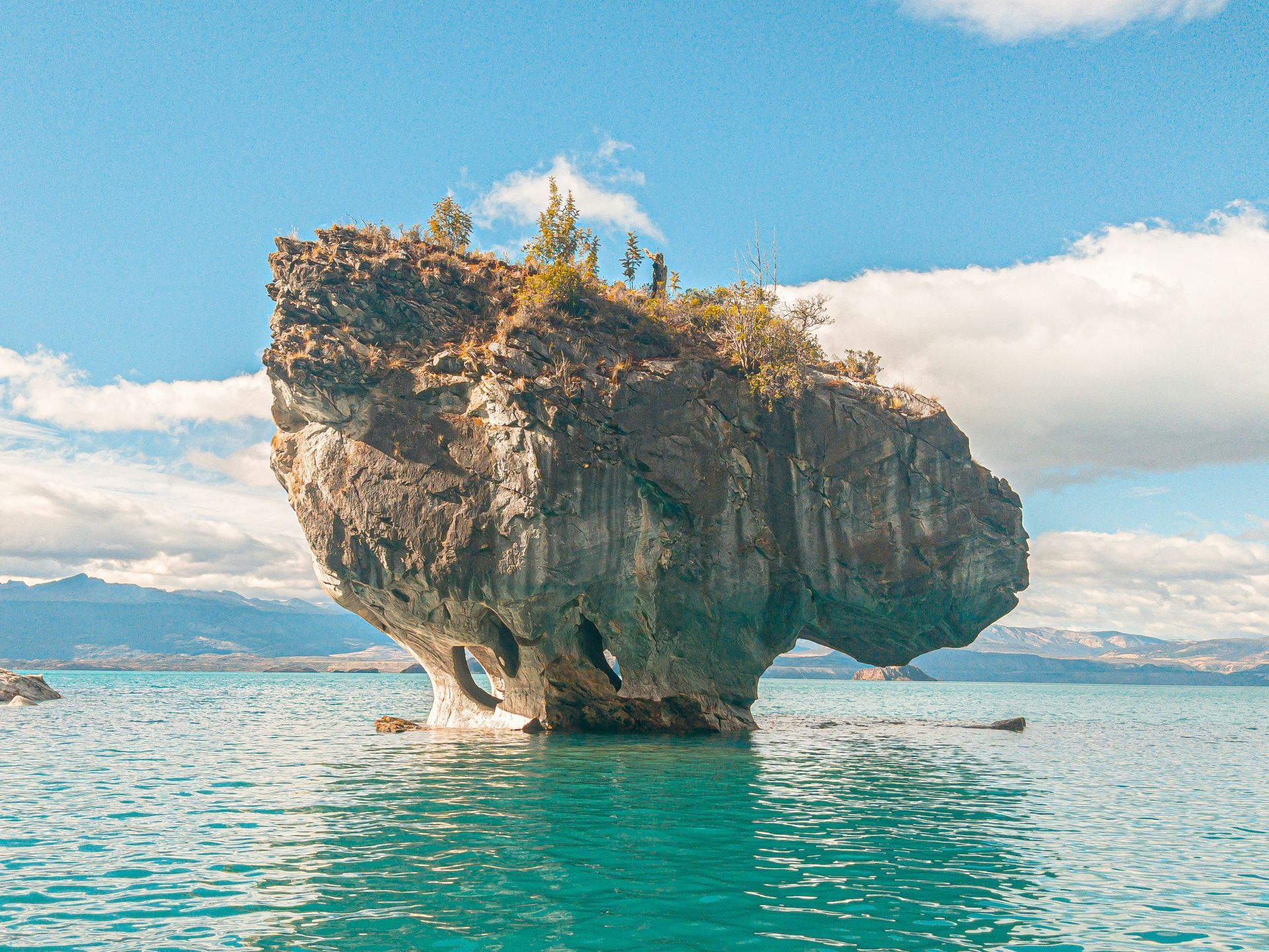 Rock formation in turquoise water, resembling a mushroom, with vegetation on top in Aysén, Chile.