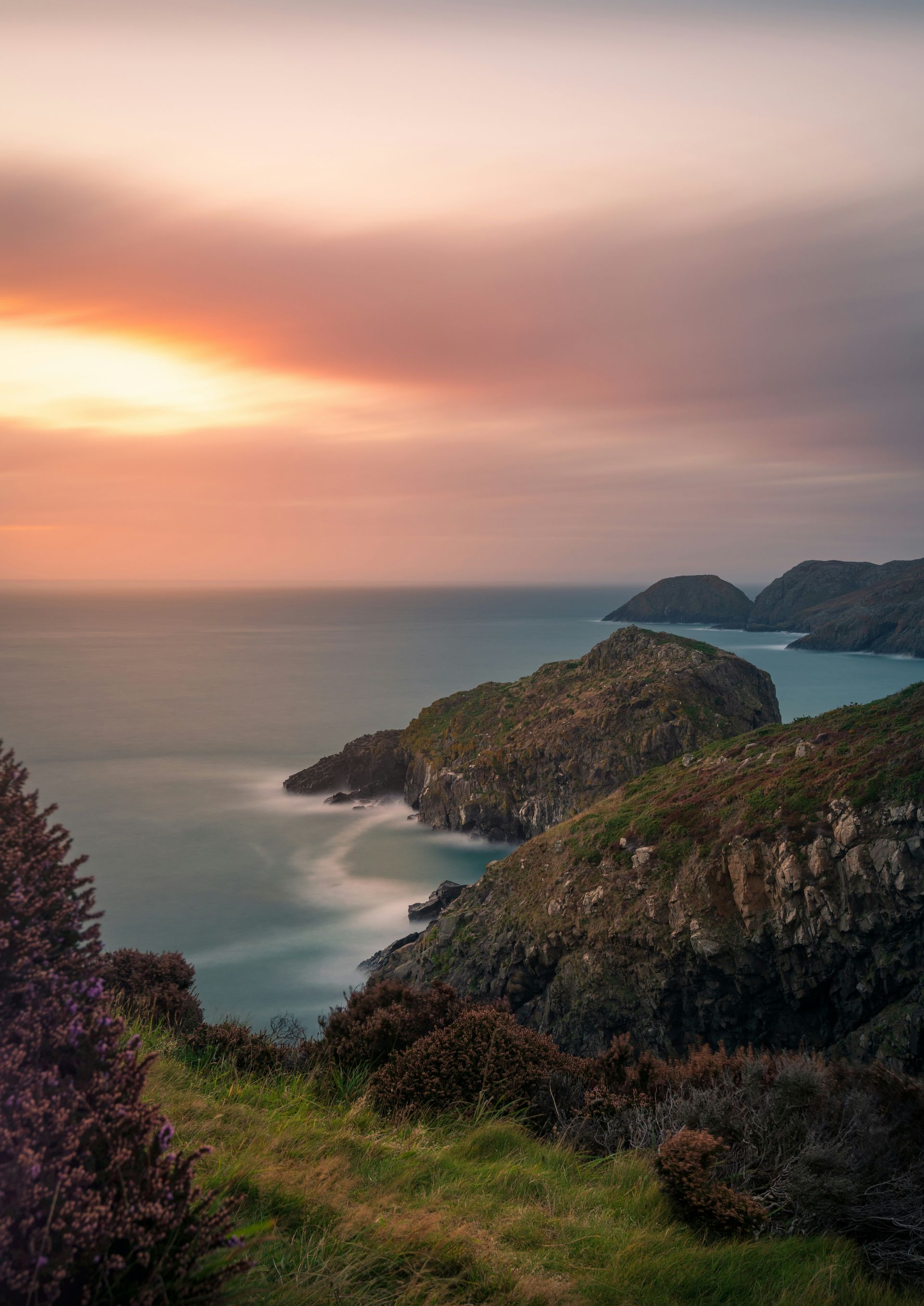 Sunset over a rugged coastline. Waves meet rocky cliffs, lush greenery below a pink and orange sky in Pembrokeshire, Wales.