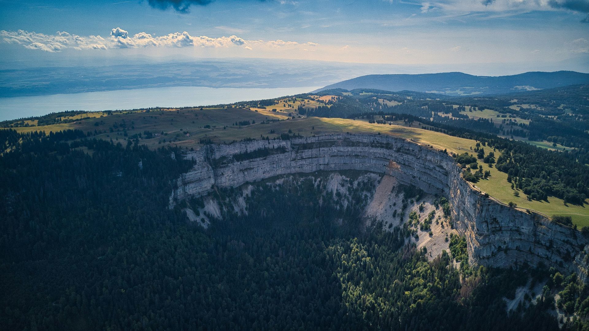 Aerial view of a Creux du Van with a large cliff face, forest, and a lake in the distance under a sunny sky in Switzerland. 