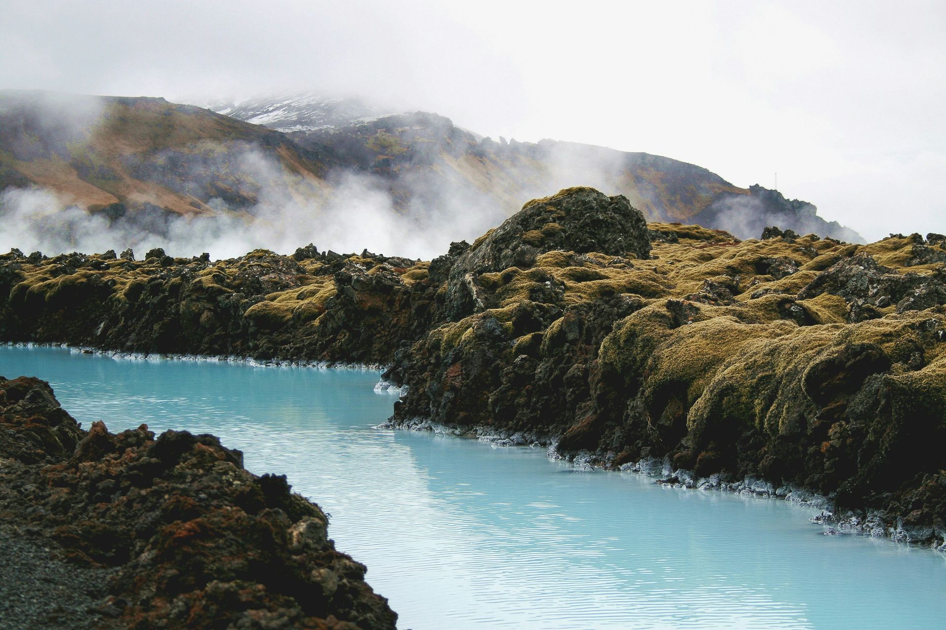 A turquoise geothermal river flows through mossy lava rock, with misty mountains in the background at Blue Lagoon, Iceland.