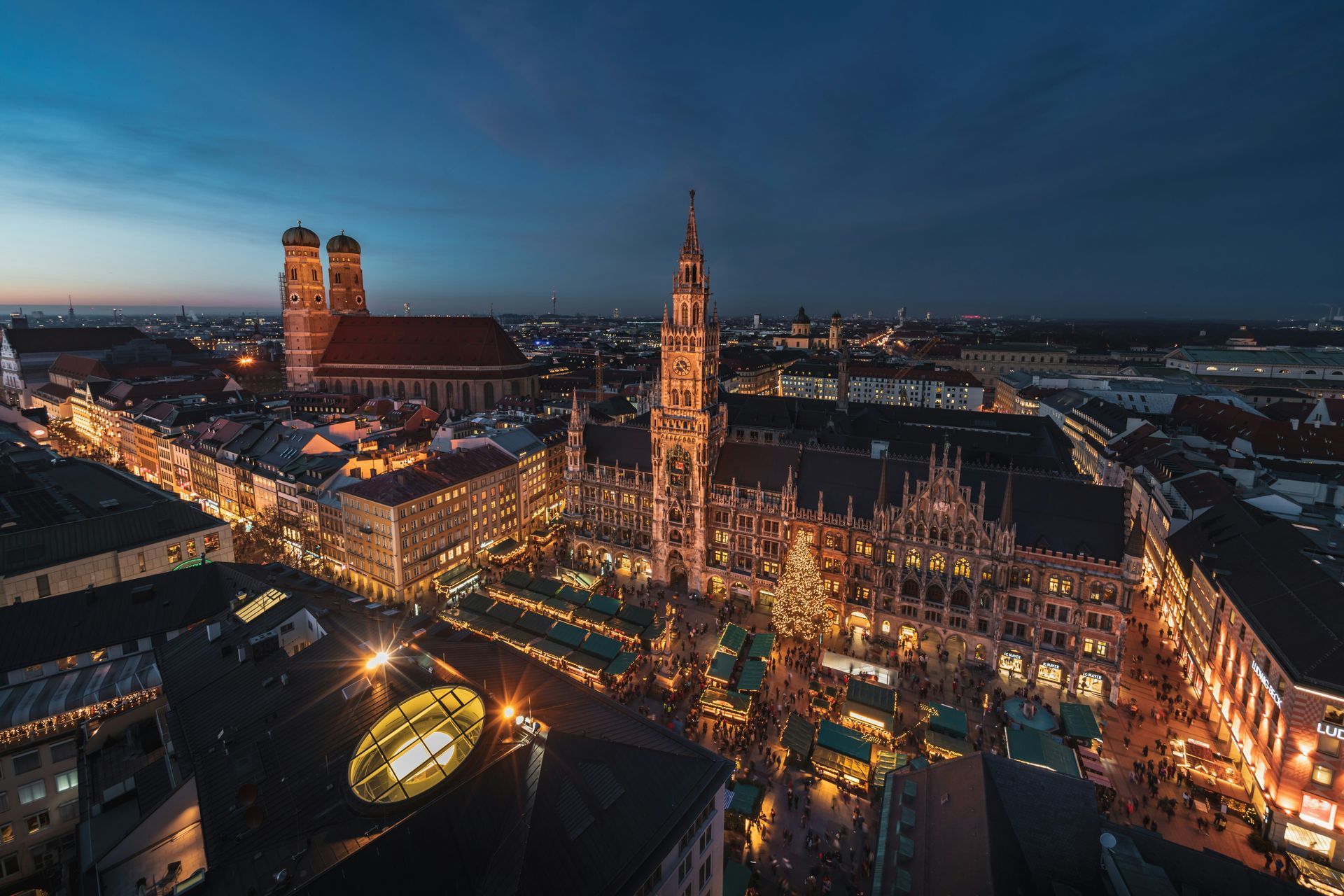 Munich cityscape at dusk, illuminated buildings, tall church spires, bustling square with market stalls and twinkling lights.