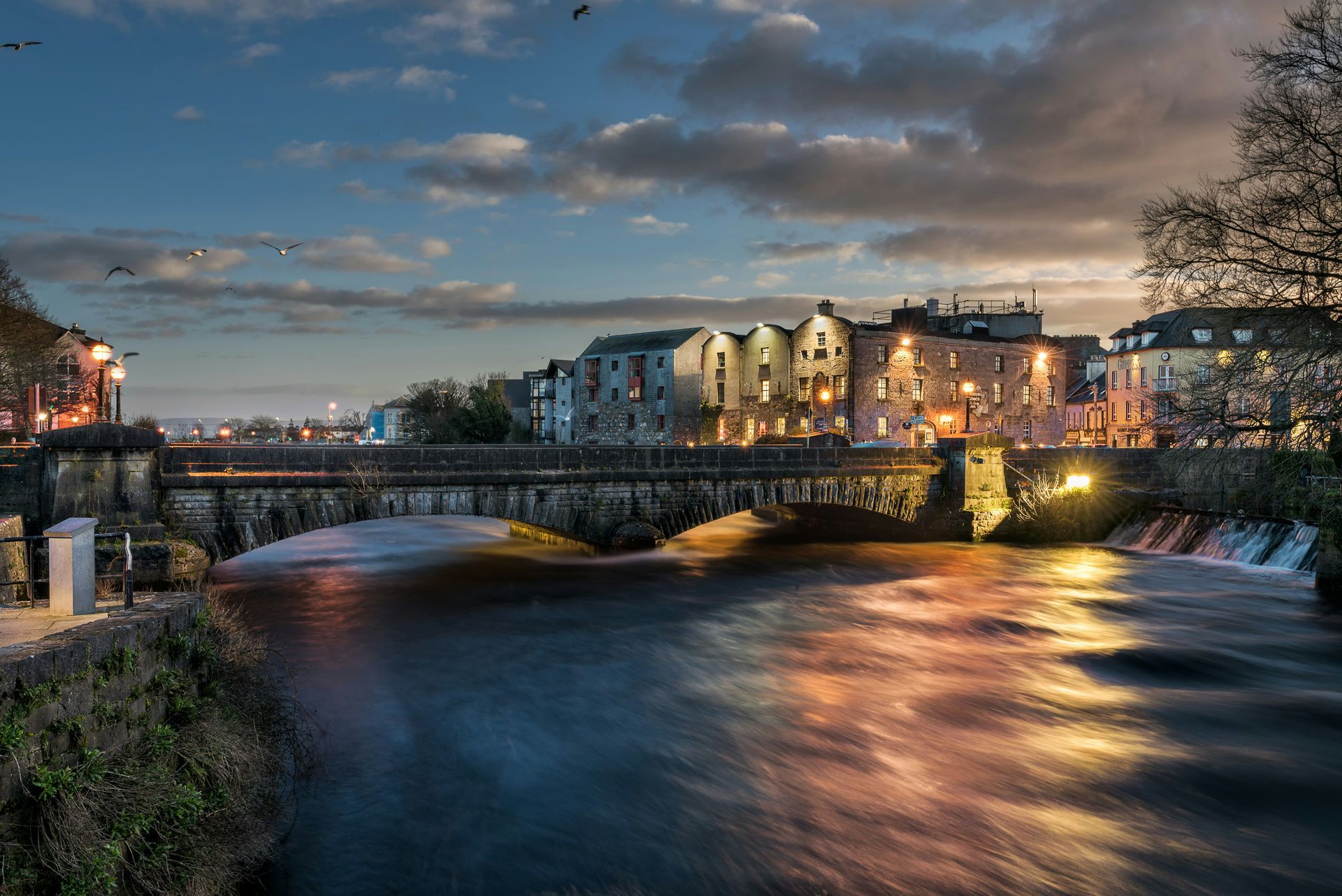 River flowing under a bridge in Galway, Ireland at dusk; buildings with lights reflect in the water.