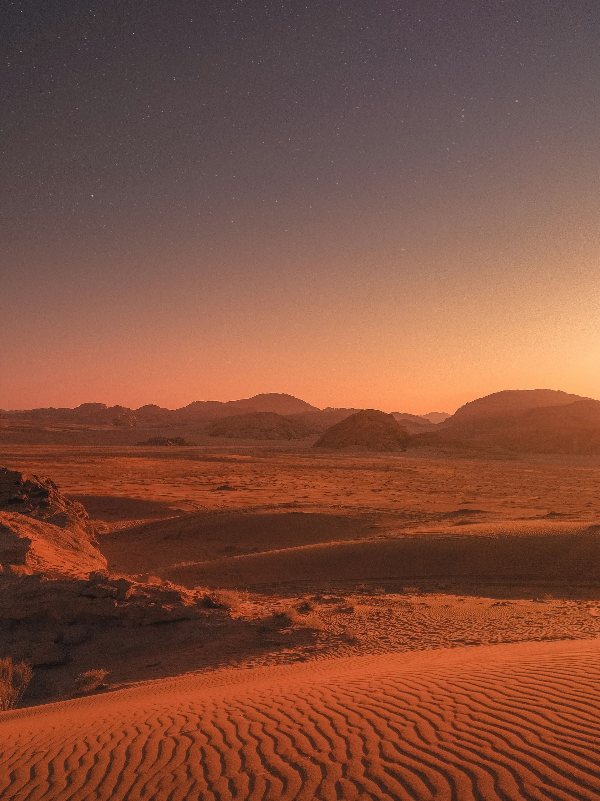 Desert landscape at dusk with a starry sky, reddish hues, and sand dunes in the foreground in Wadi Rum, Jordan.
