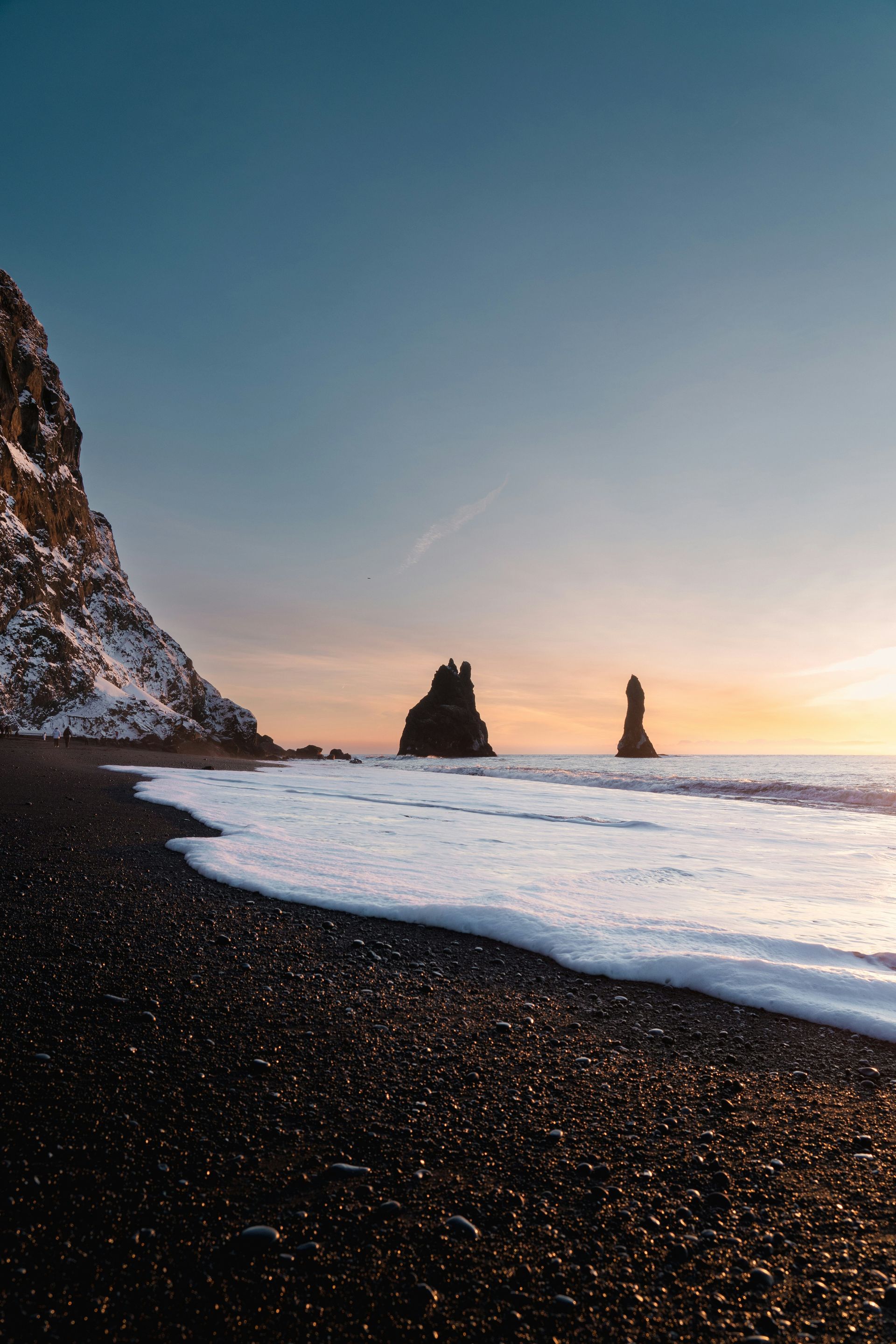 Reynisfjara Beach is a black sand beach with sea foam, two rock formations, mountain, and sunset sky in Iceland.