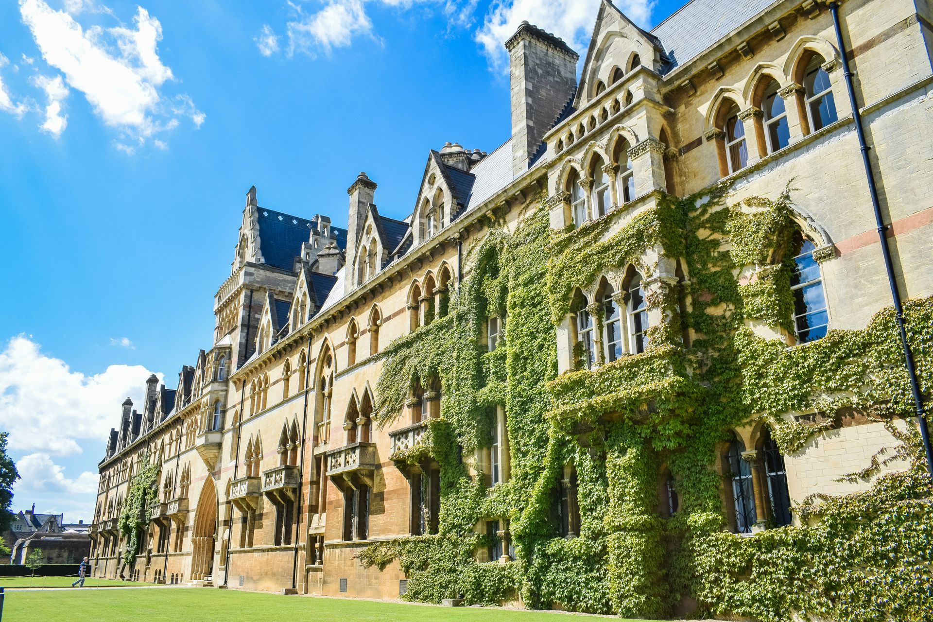 Ivy-covered sandstone building, likely a college, with arched windows under a bright blue sky at Oxford University, England.
