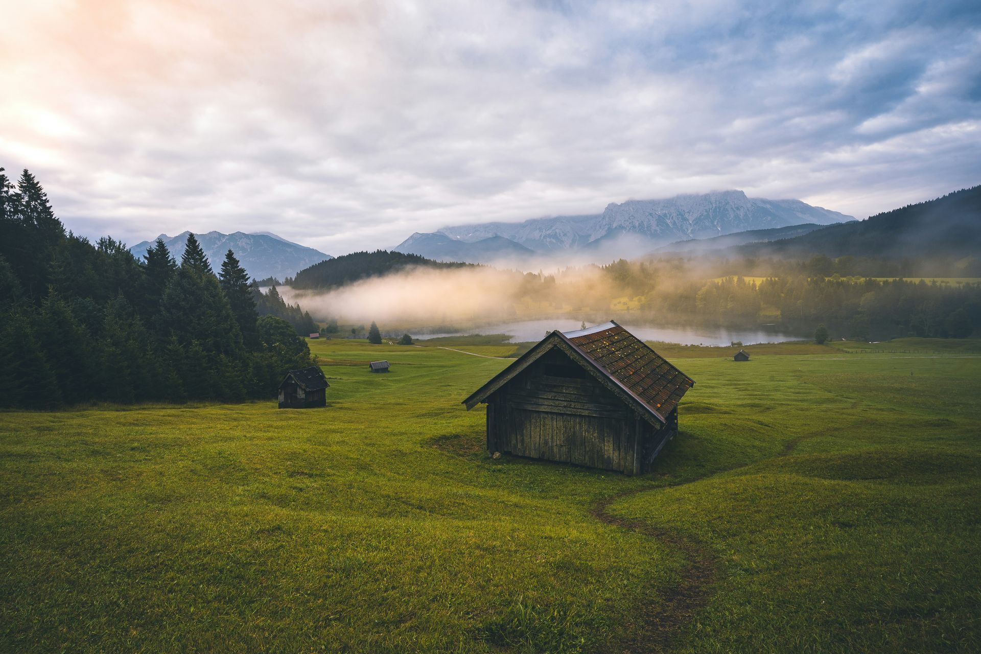 Wooden cabin in a green field with a misty lake and mountains in the background under a cloudy sky in Krün, Germany.