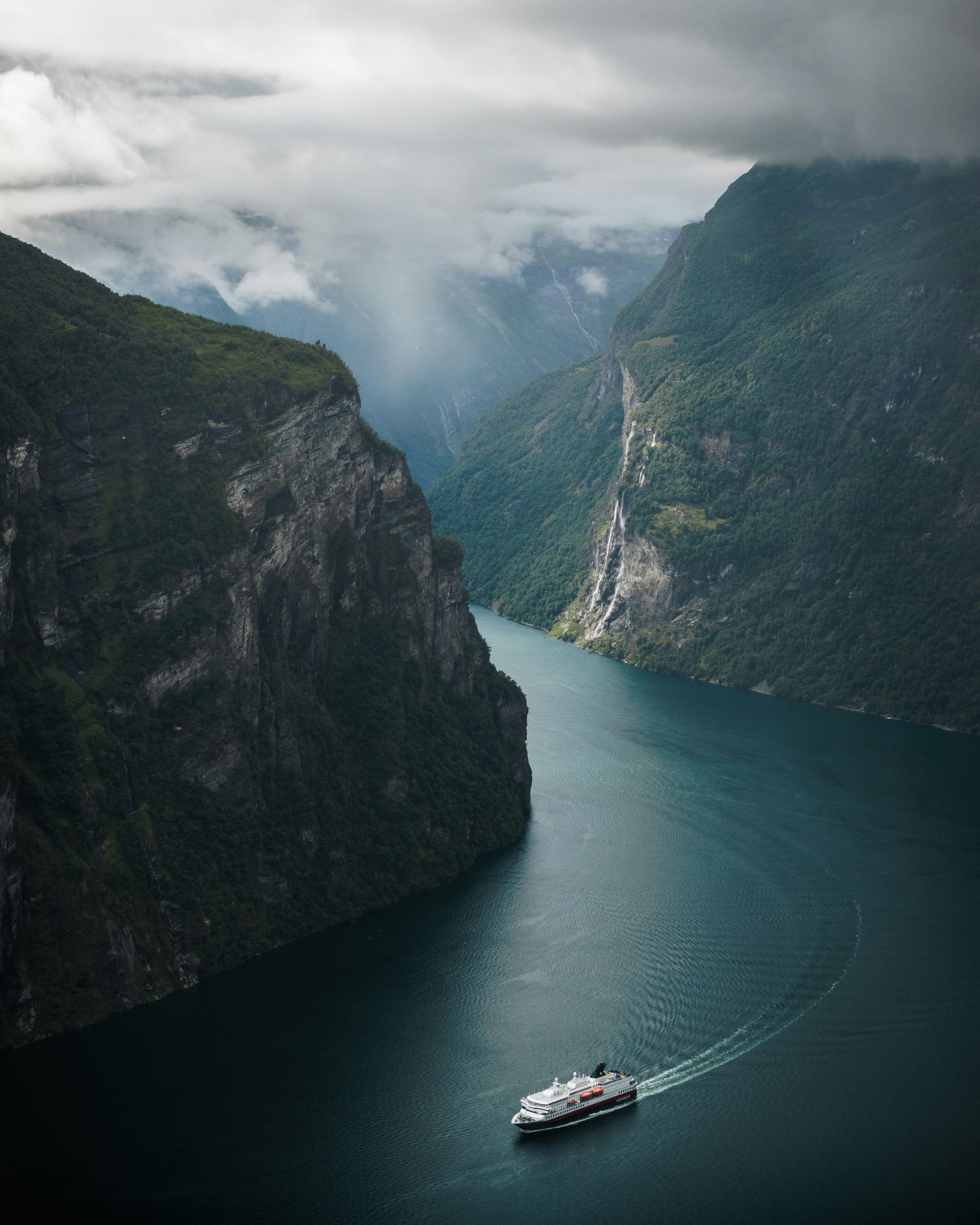 Geirangerfjord with cruise ship, flanked by steep, green cliffs, waterfall, and an overcast sky in Norway.