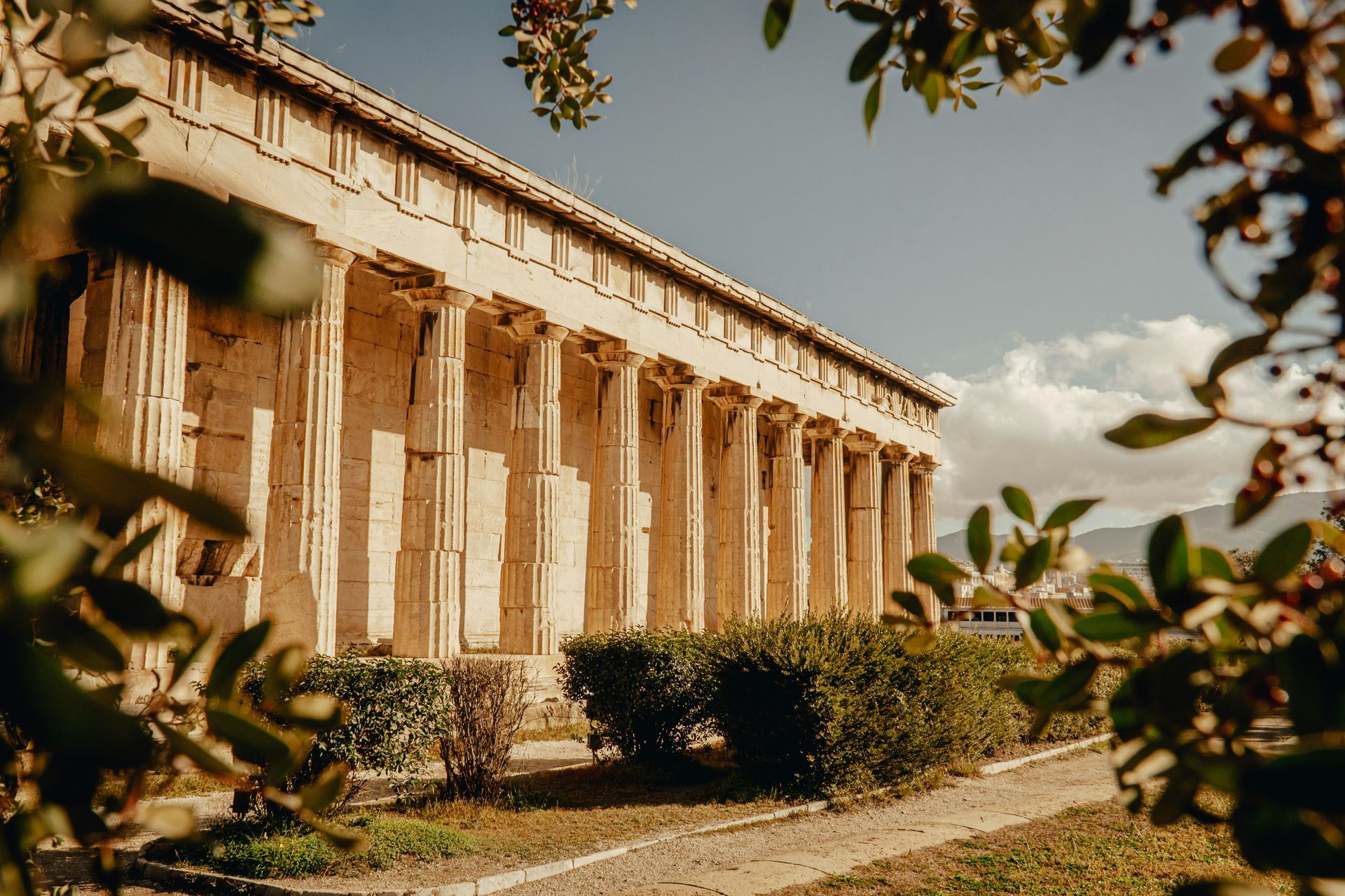 A large building with columns and a roof is surrounded by trees and bushes in Greece.