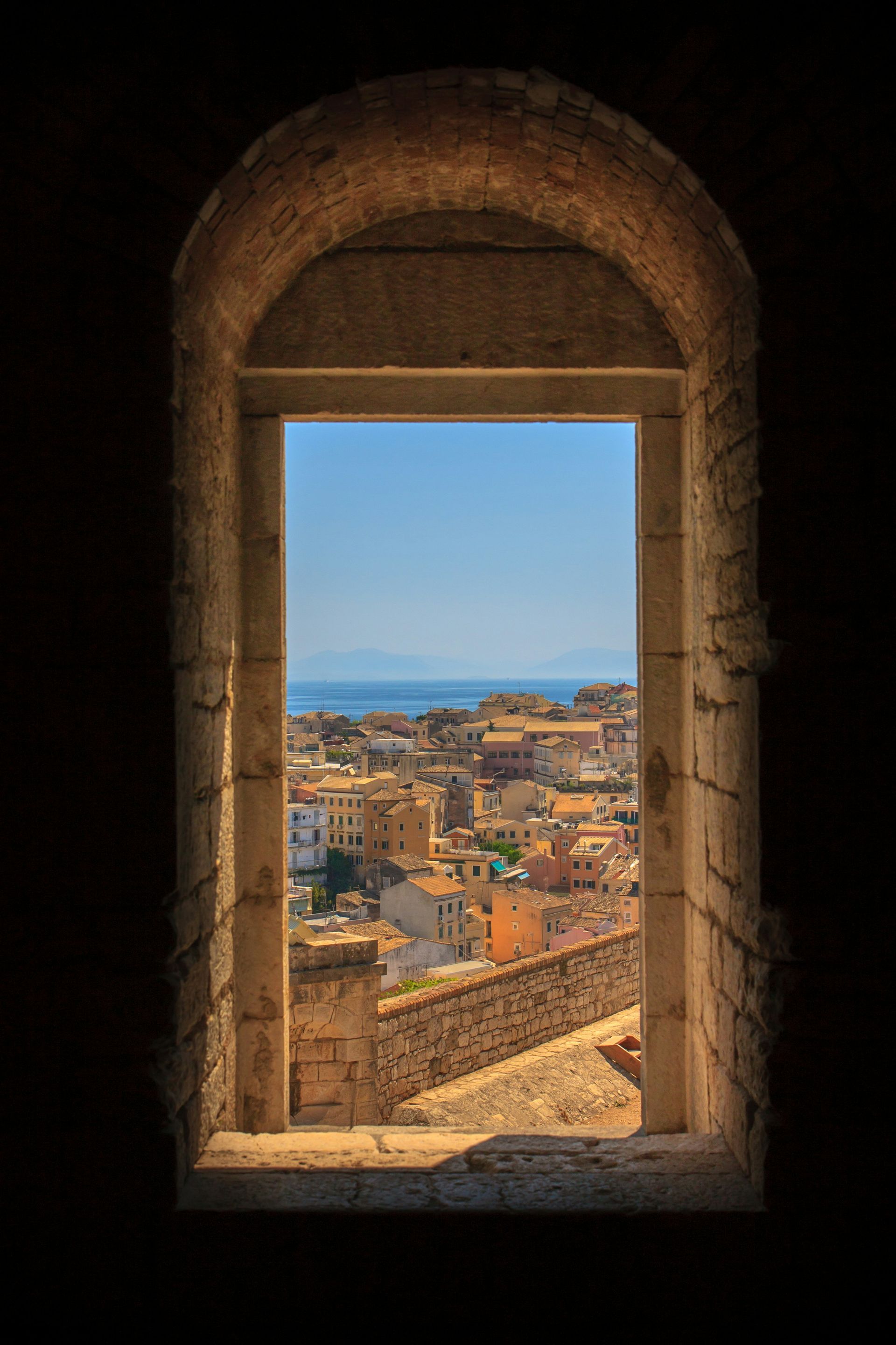 A window with a view of a city and the ocean in Corfu, Greece.