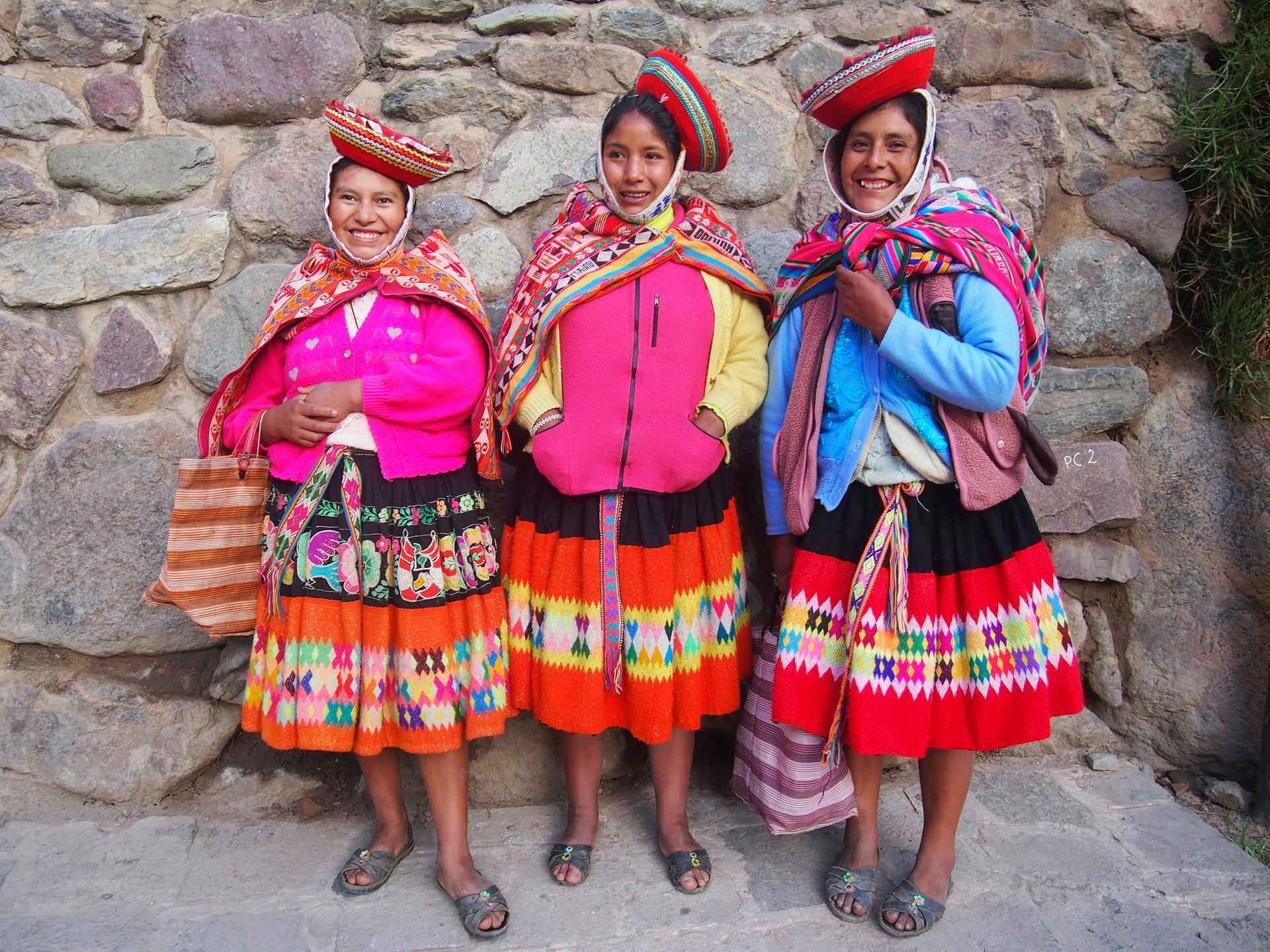 Three women in traditional Peruvian clothing are standing in front of a stone wall in Peru.