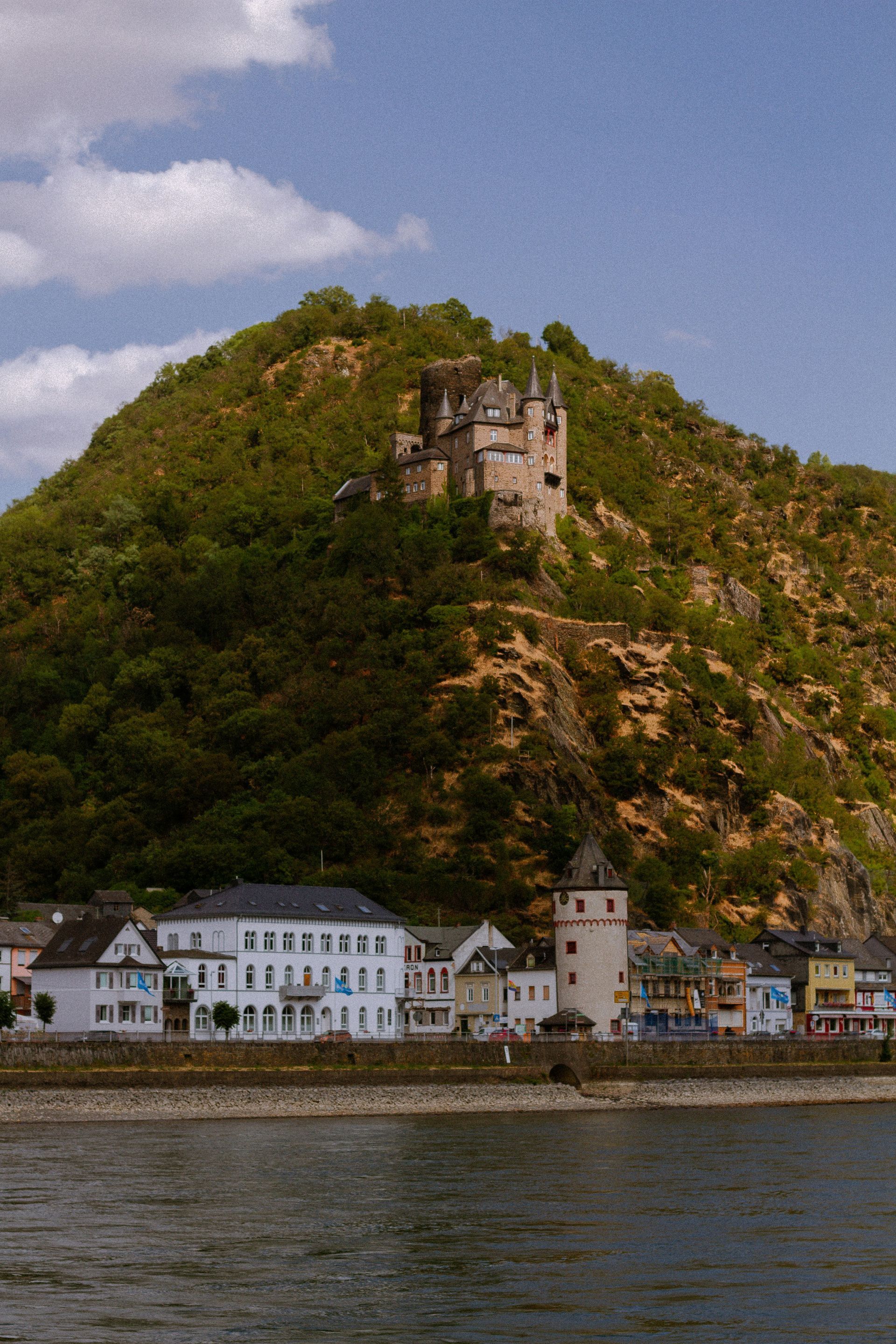 A castle perched on a lush green hillside overlooks the town of Bacharach on the Rhine River in Germany.