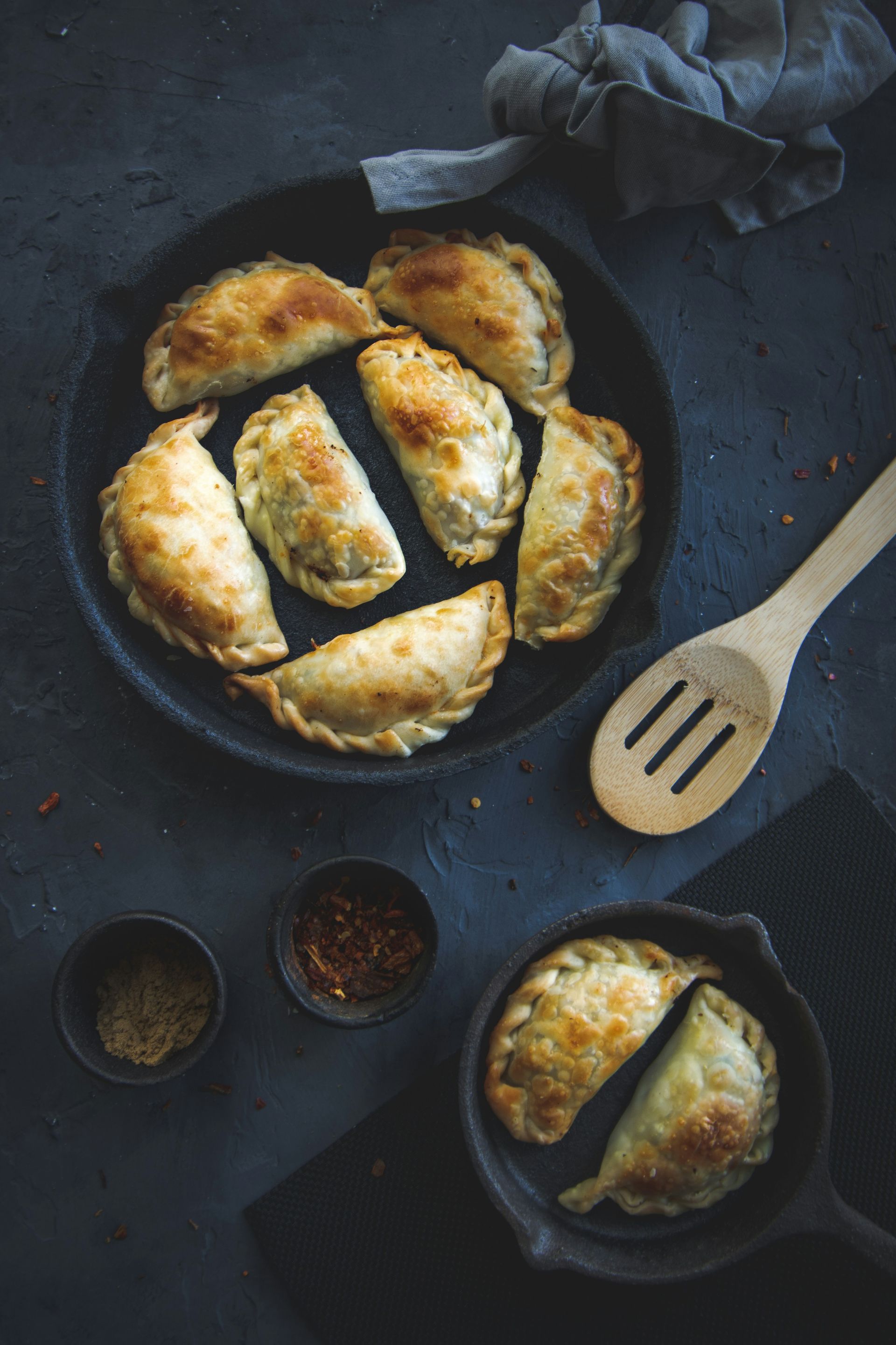 Golden-brown empanadas on a black plate with spice bowls, a wooden spoon, and a dark background in Argentina. 