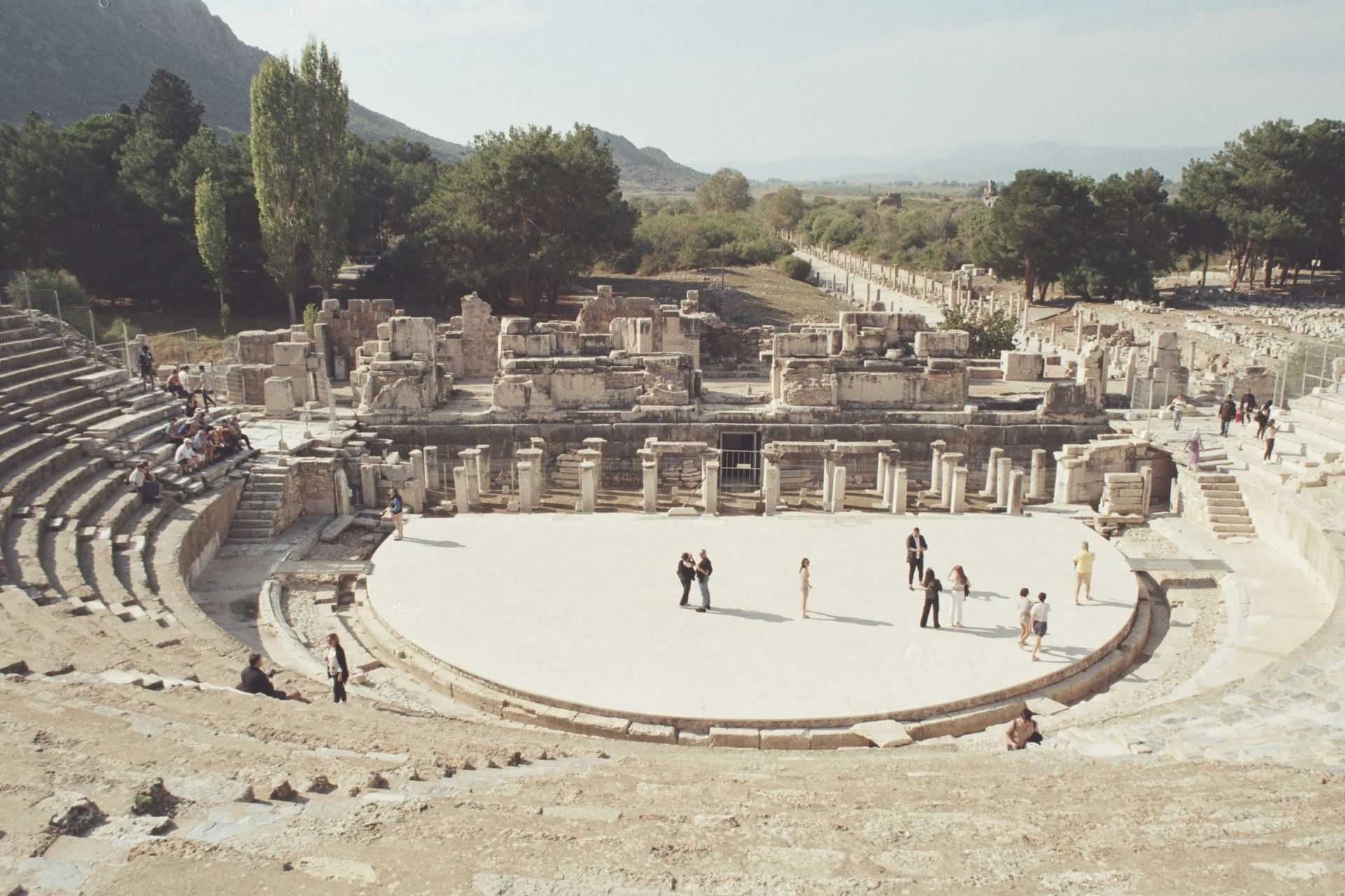 Ruins of an ancient Roman theater in Ephesus, Turkey. People stand on stage. Mountains and trees in the background.