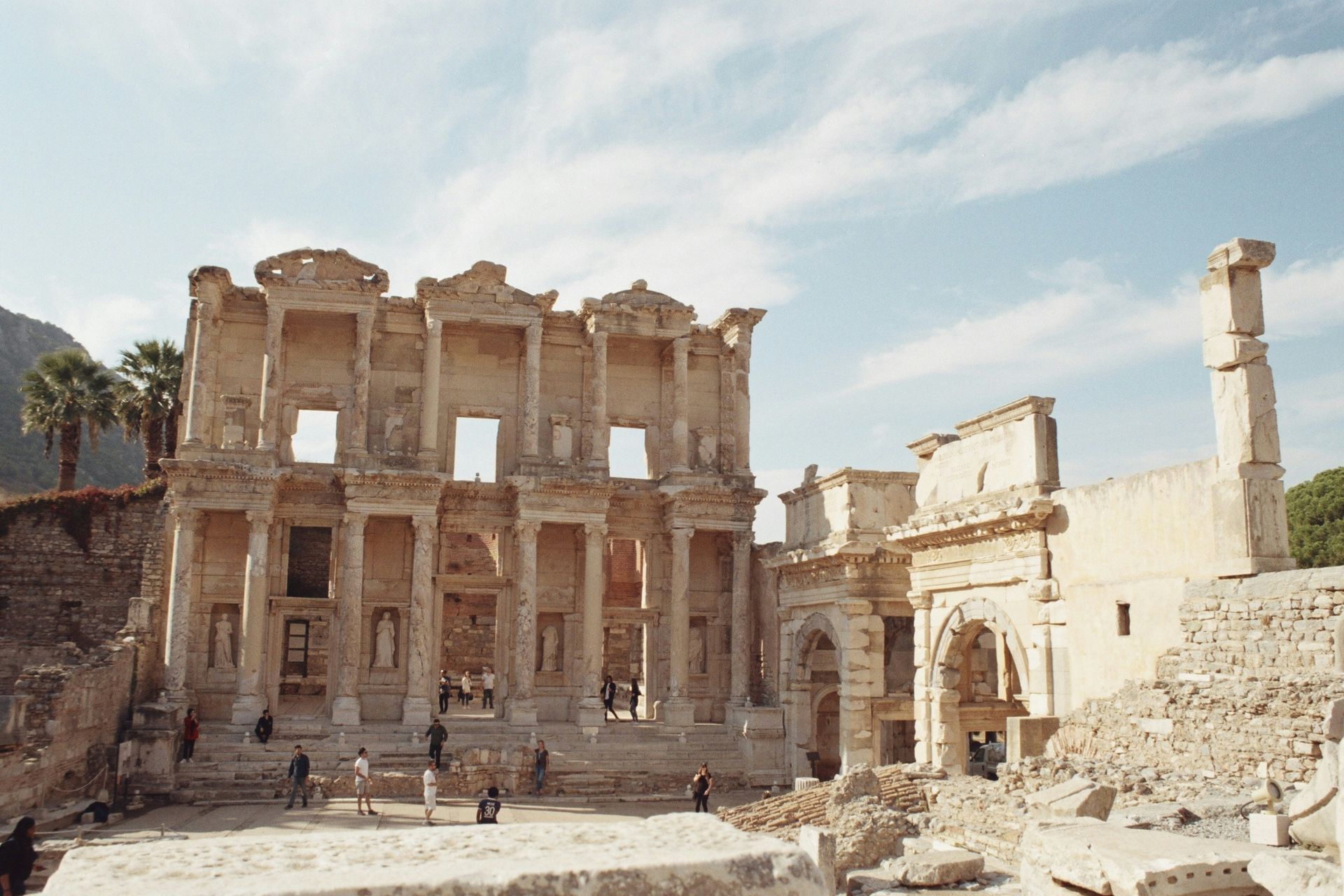 Ruins of the Library of Celsus in Ephesus, Turkey, with columns, arched doorways, and tourists on a sunny day.