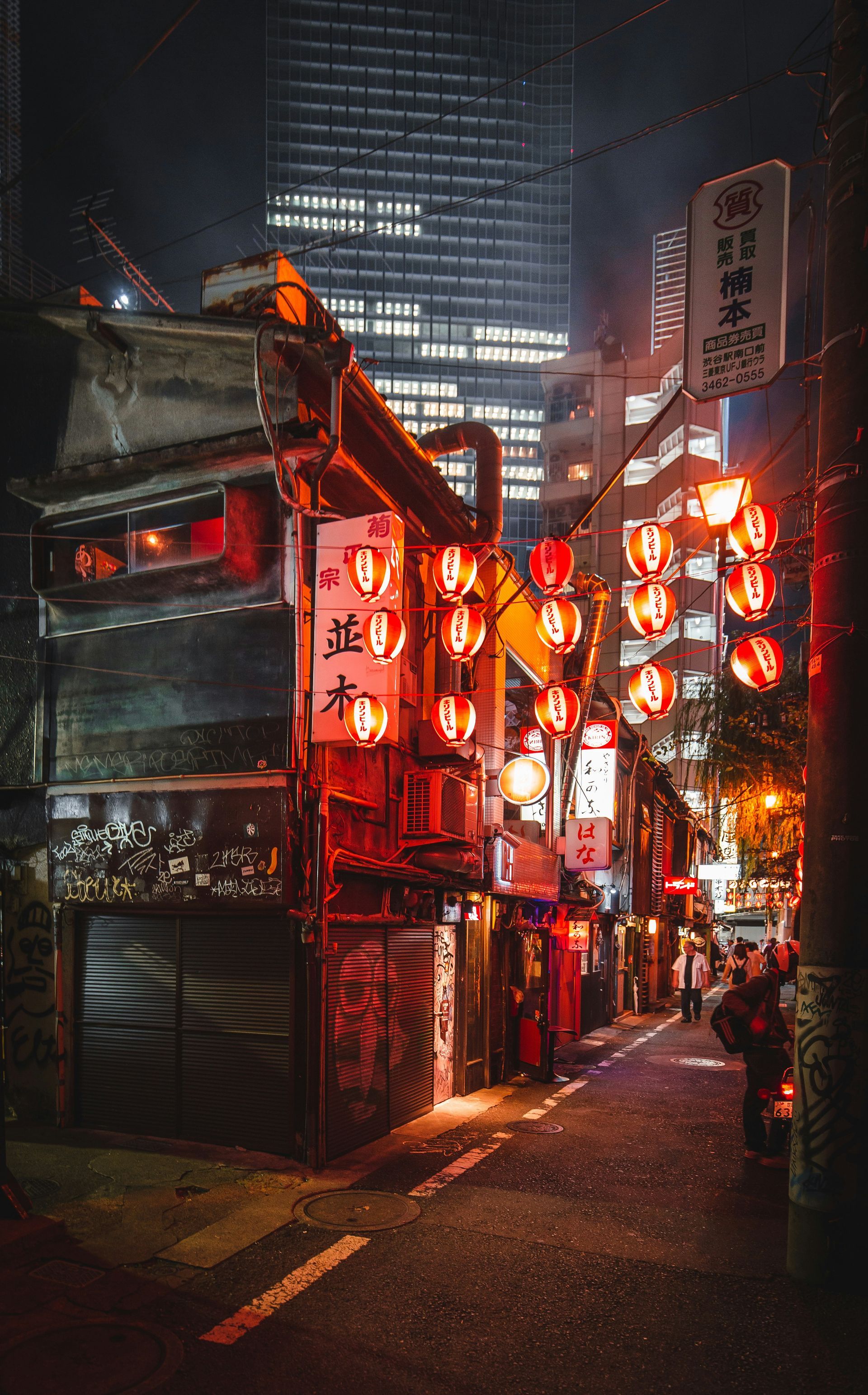 Narrow Tokyo alley at night, lined with red lanterns and illuminated signs, skyscraper in background.