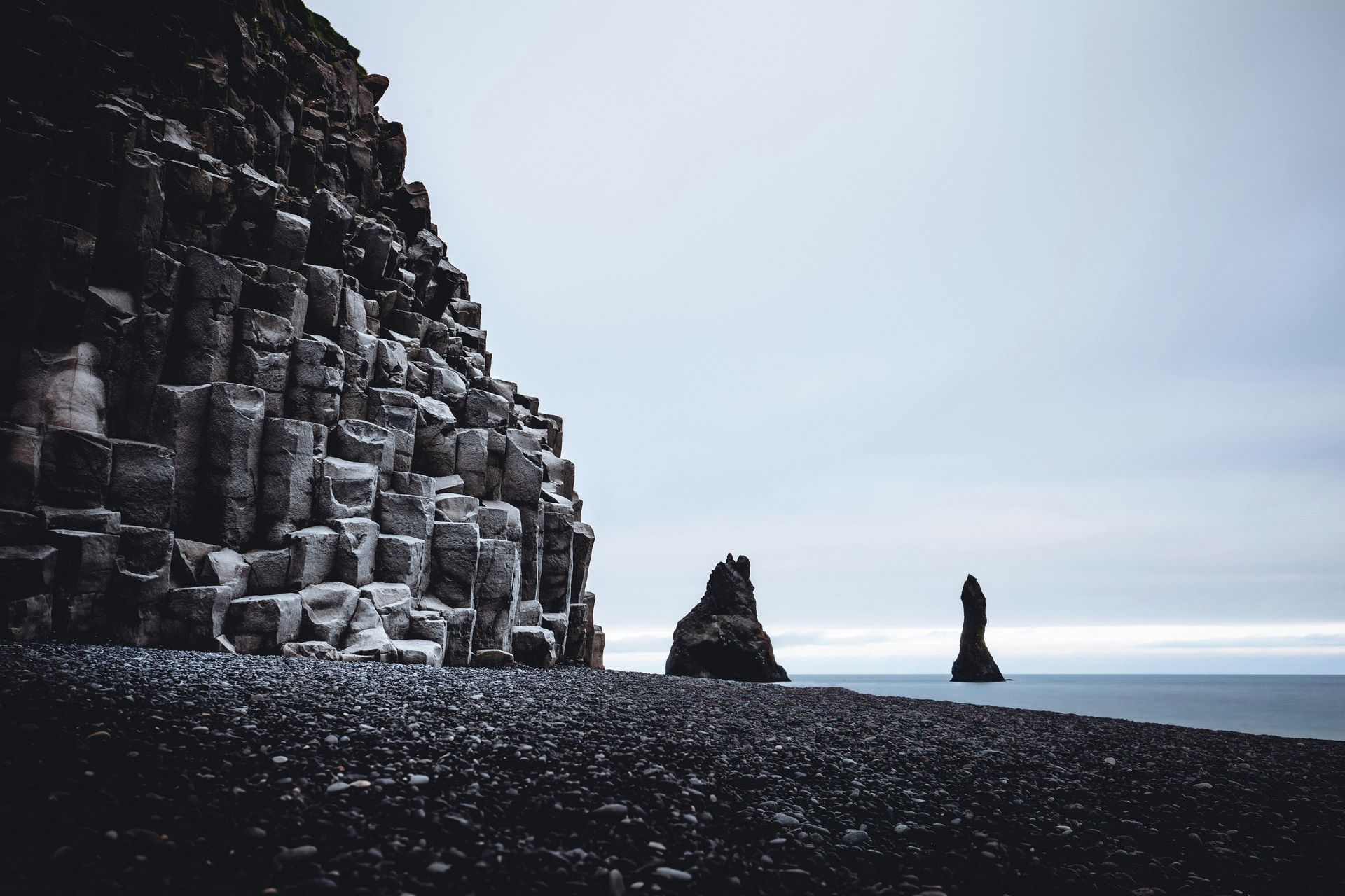 Black basalt cliffs and rock formations on a dark beach against a gray sky and ocean at Reynisfjara Beach, Iceland.