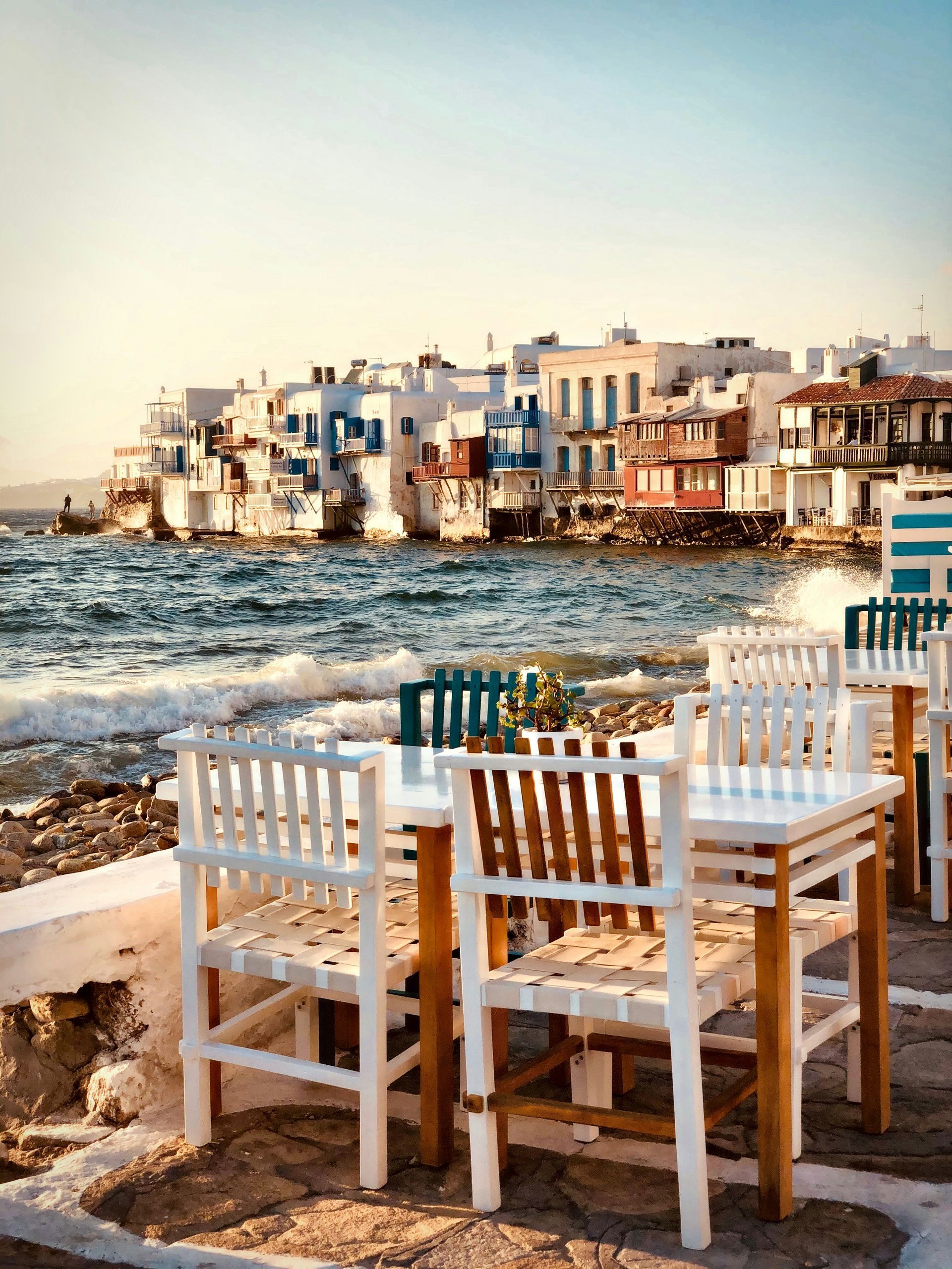 Tables and chairs on a beach with a city in the background in Mykonos, Greece.